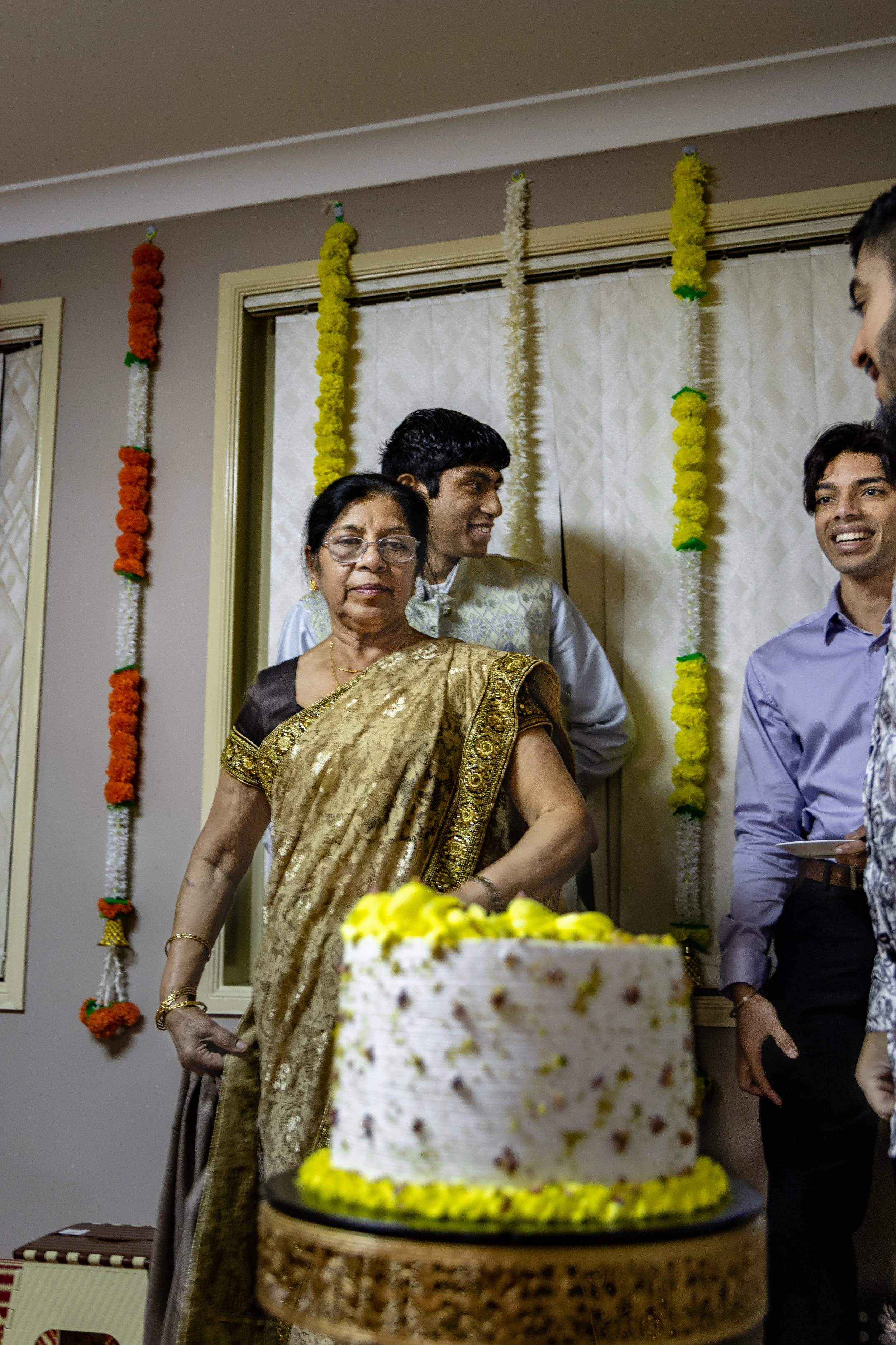 A group of people celebrating around a birthday cake decorated with yellow flowers, with colorful flower garlands hanging on the wall in the background.