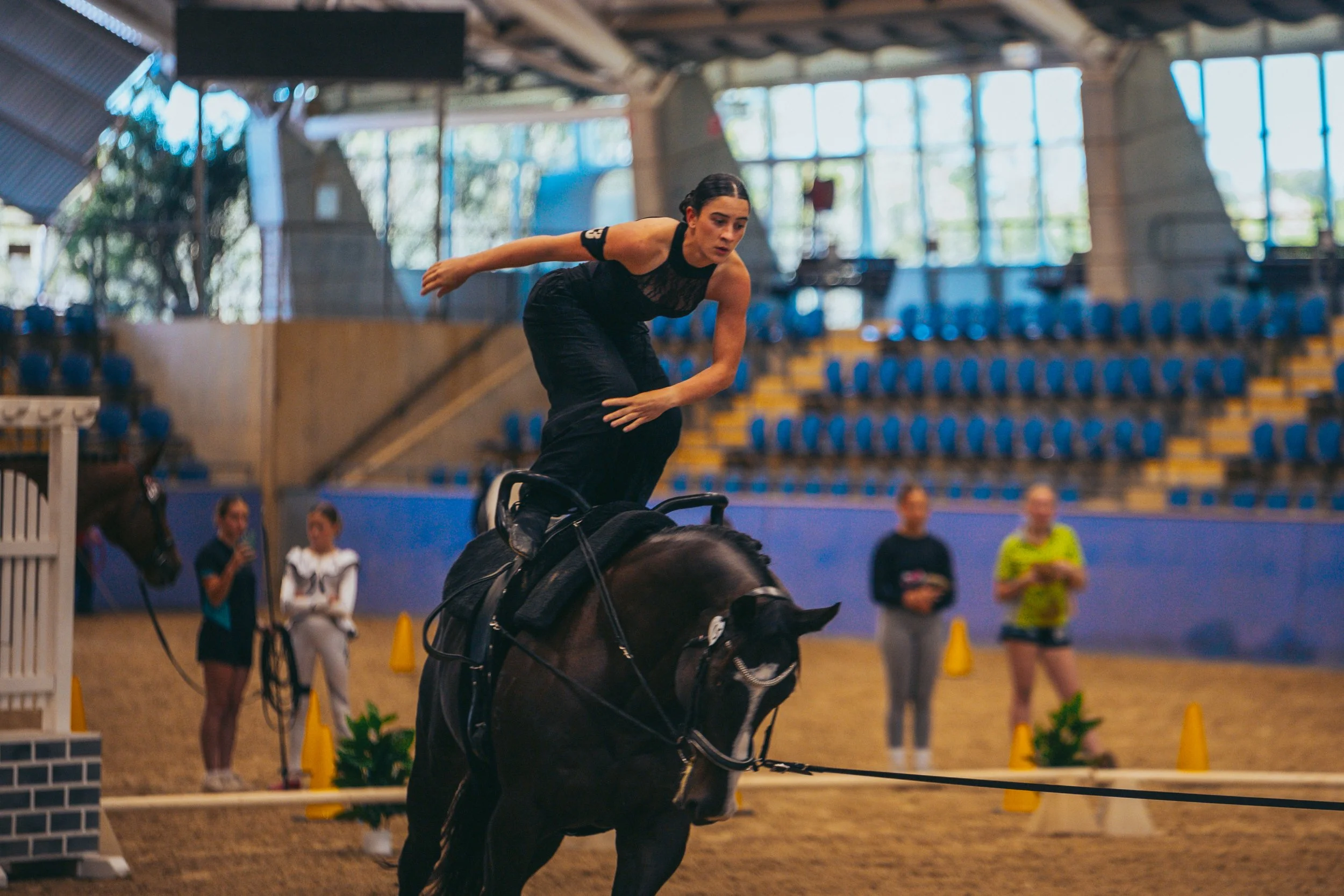 A female equestrian performs a stunt on a black horse inside an indoor riding arena with seating and large windows in the background.