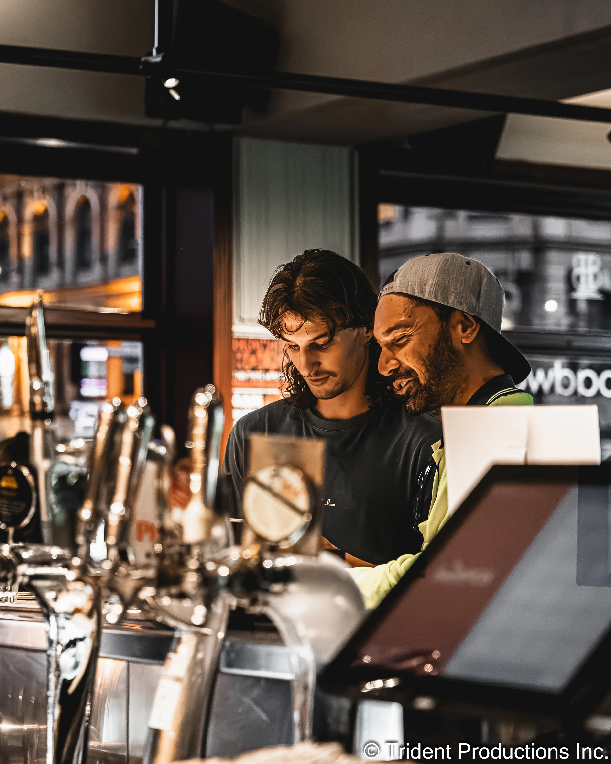Two male bartenders working behind a bar, focused on a task, in a dimly lit bar or pub with beer taps and a digital ordering screen.