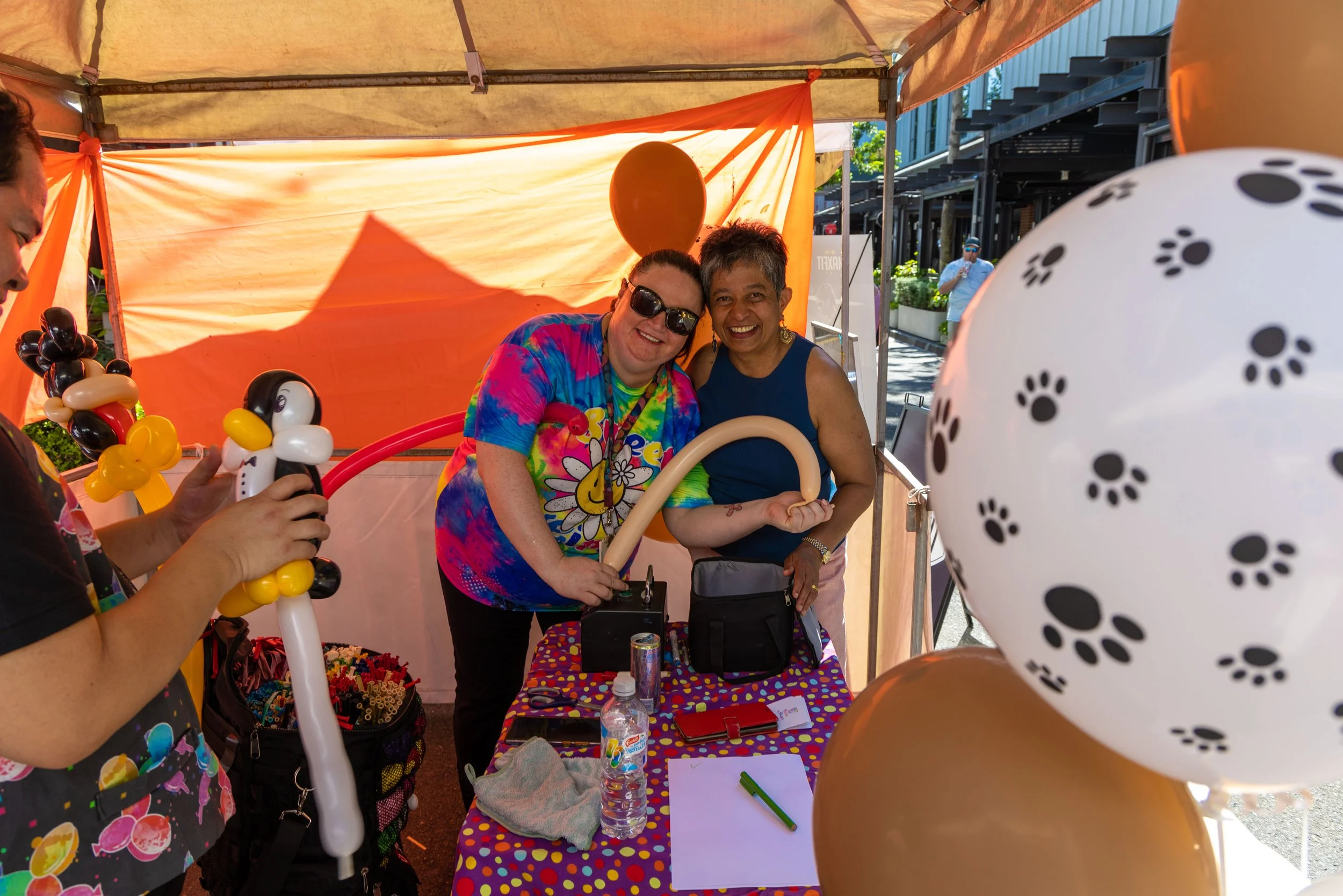 Two smiling women in a booth with colorful balloons, one holding a balloon animal, at an outdoor event under an orange canopy.