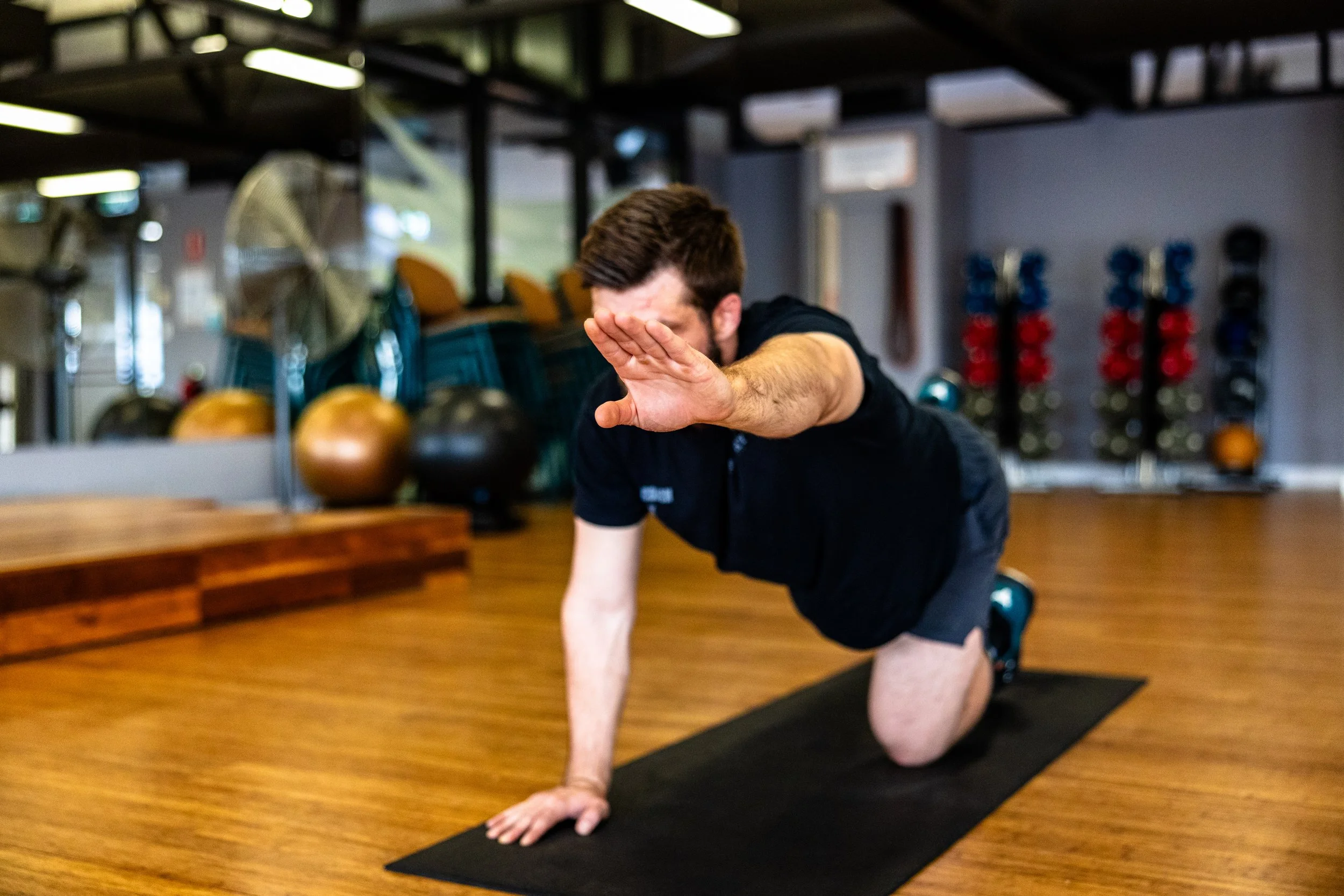 A man in a black shirt and shorts doing a yoga pose on a black mat in a gym, with one arm extended forward and the other supporting him on the floor.