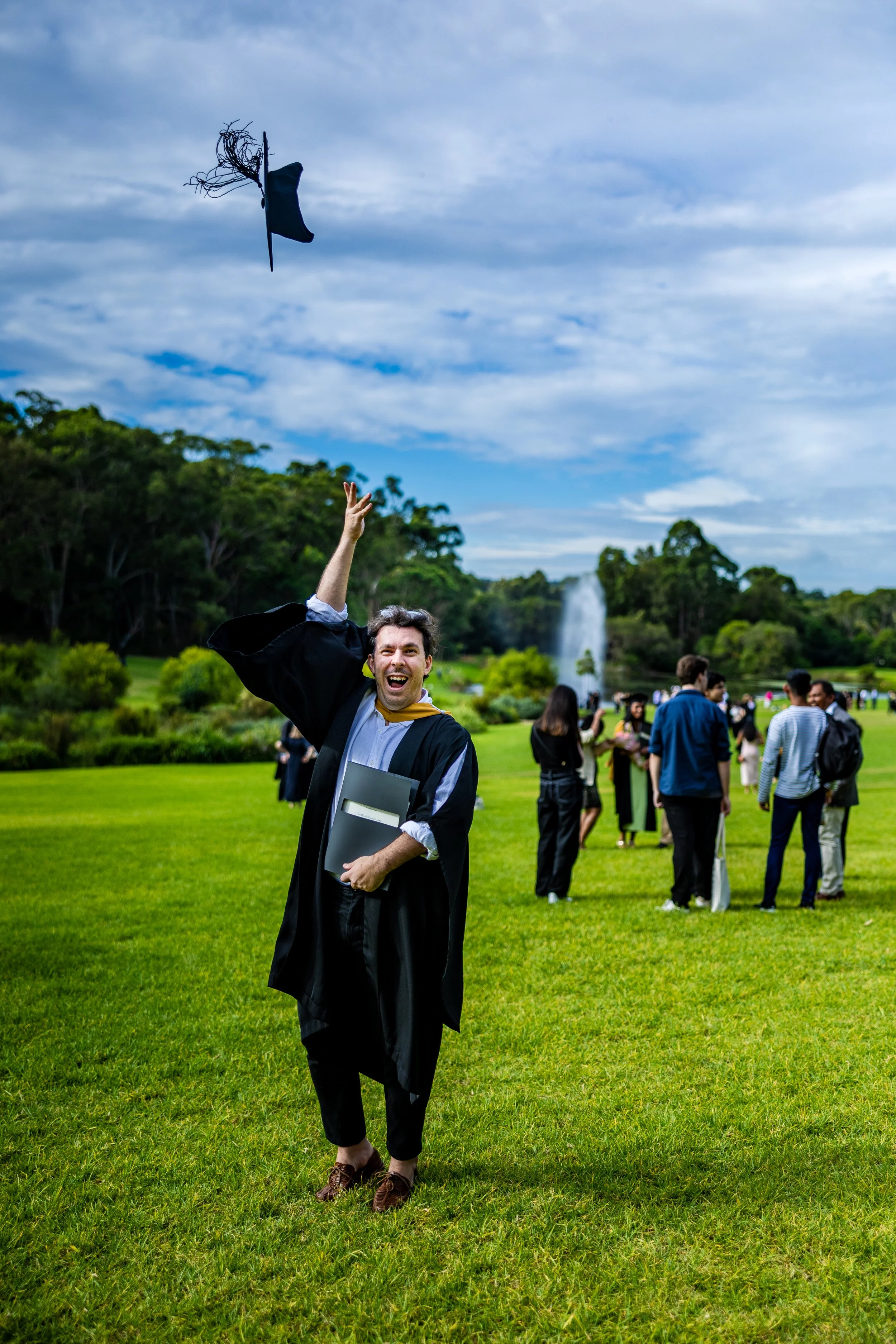 A graduate in a black gown and cap throwing their cap in the air on a grassy field with a fountain in the background, celebrating graduation.