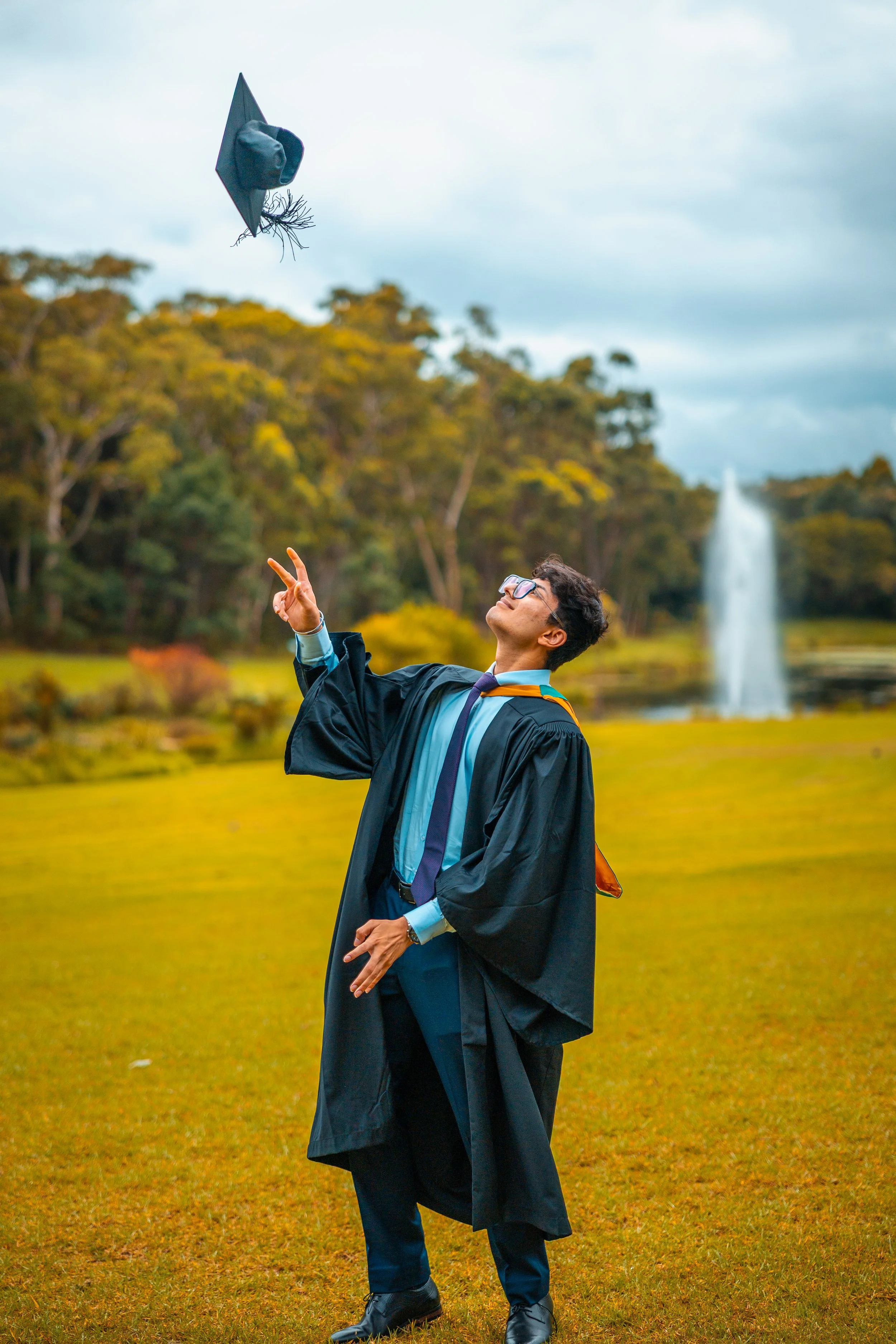 A young man in graduation regalia tossing his cap in the air outdoors near a lake with a fountain, trees, and a cloudy sky in the background.