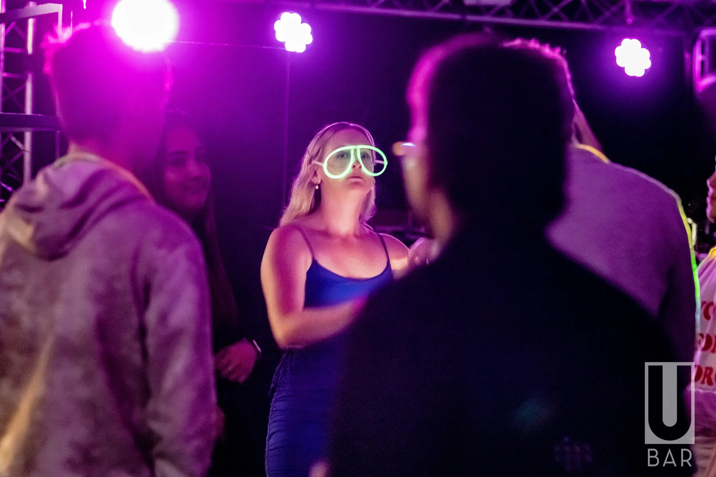 People at a nightclub with purple lighting; one woman is wearing glowing neon glasses and talking to others.