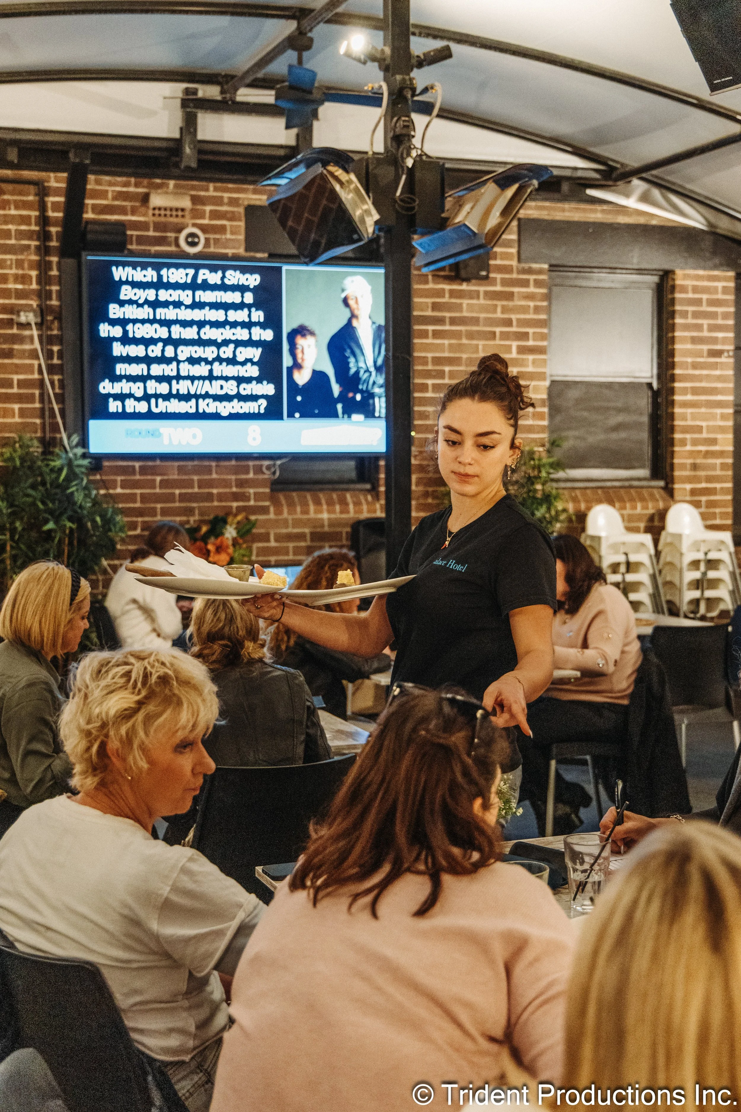 A woman in a black shirt is serving food to a group of people seated at tables in a restaurant or event space with exposed brick walls and a large television screen in the background displaying a trivia question.