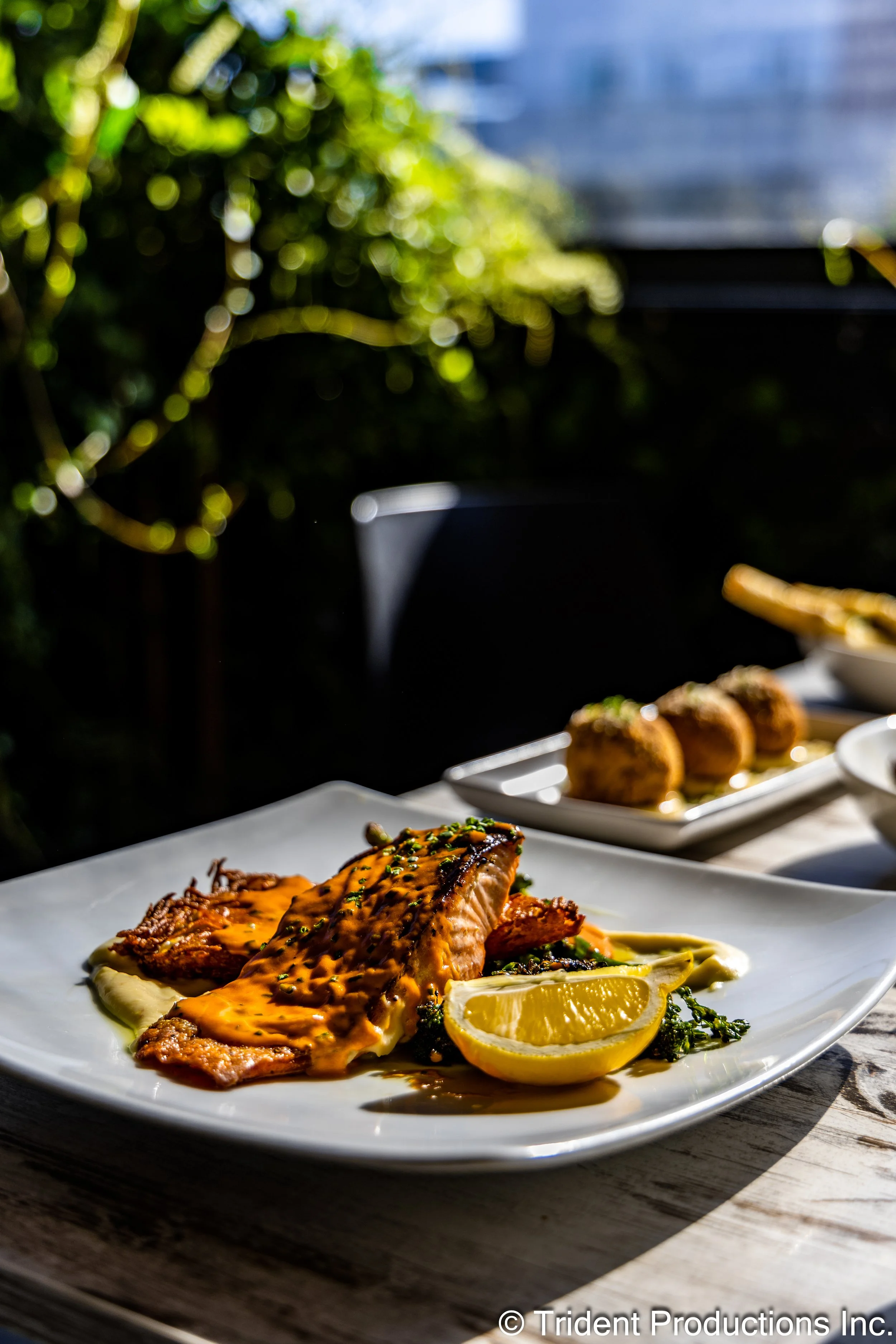 A plate of grilled fish topped with sauce, served with lemon wedges, with other dishes in the background on an outdoor table.