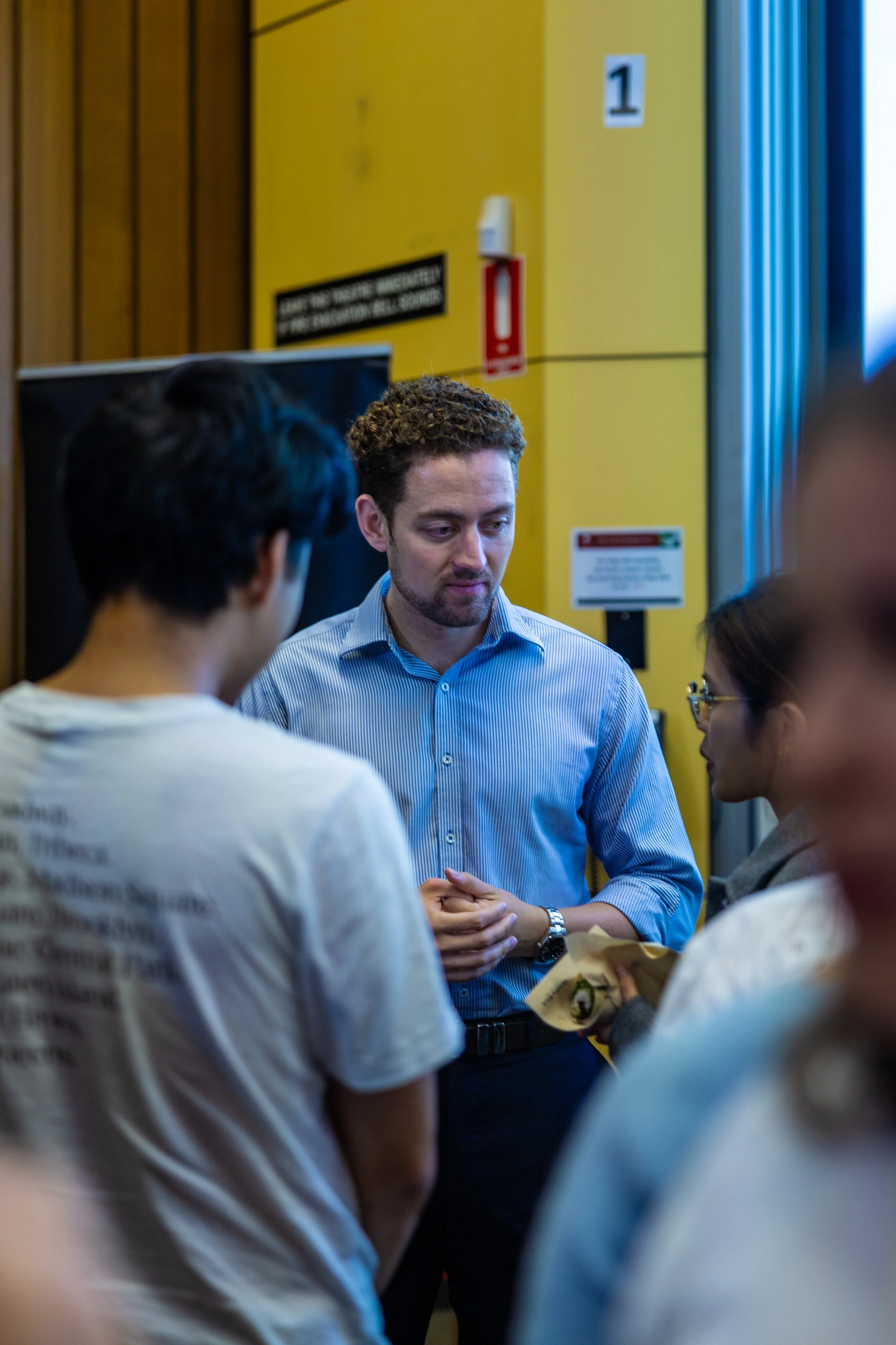 A man with curly hair and a blue striped shirt speaking to a group of people indoors.