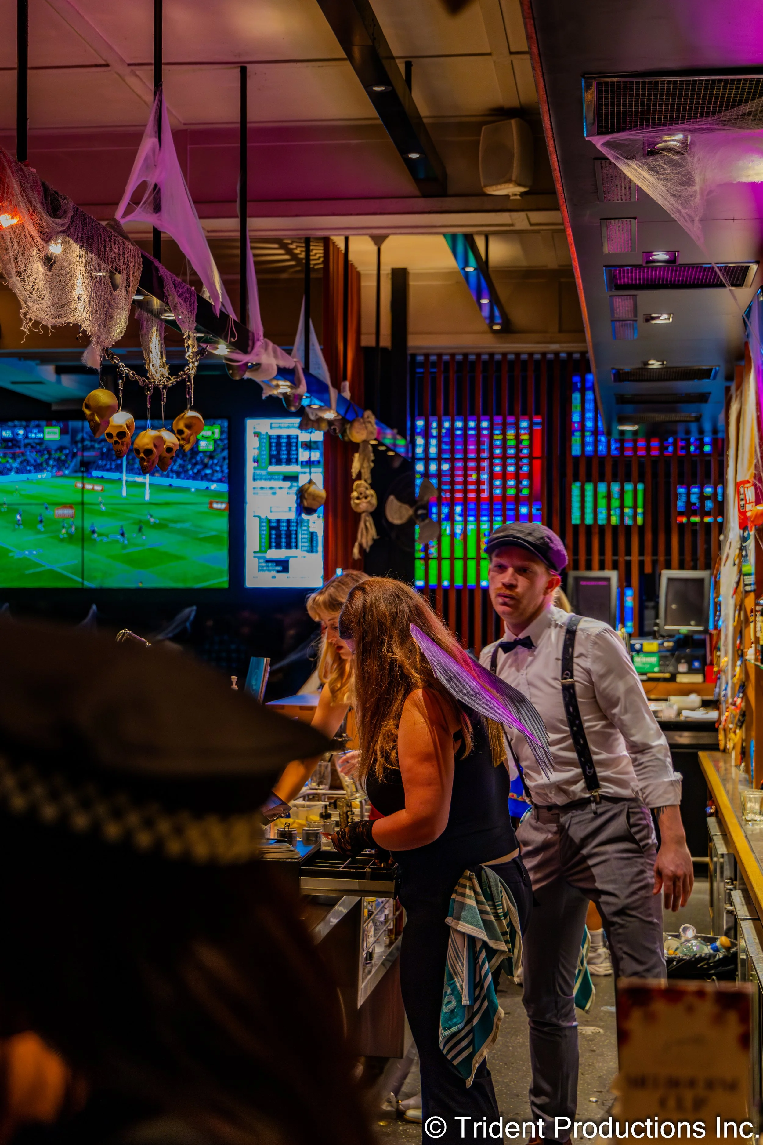 People dressed in Halloween costumes at a bar decorated with cobwebs and skull-shaped ornaments, with a TV screen displaying a sports game in the background.
