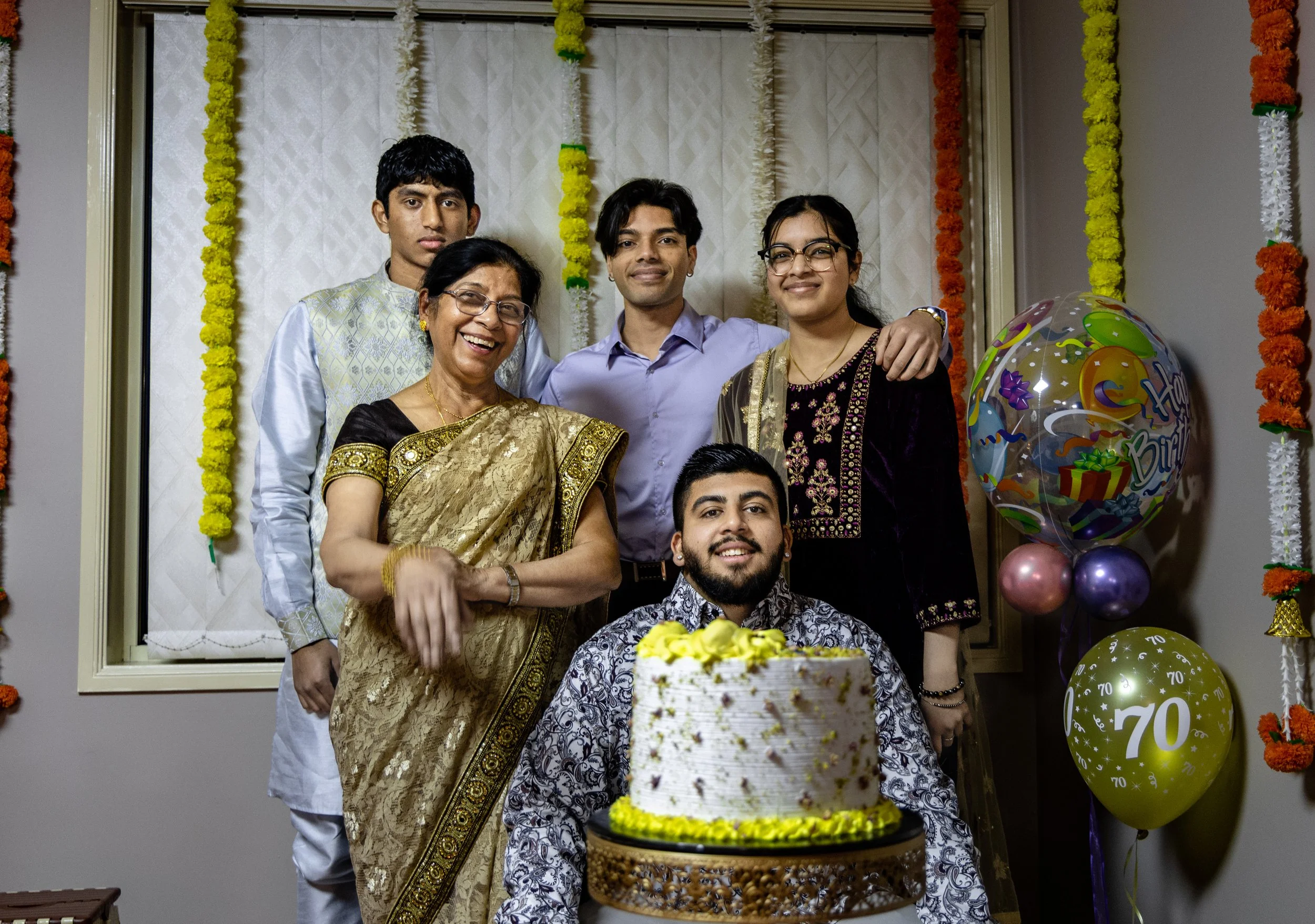 Group of six people celebrating a 70th birthday, with a cake, balloons, and festive decorations in a decorated room.