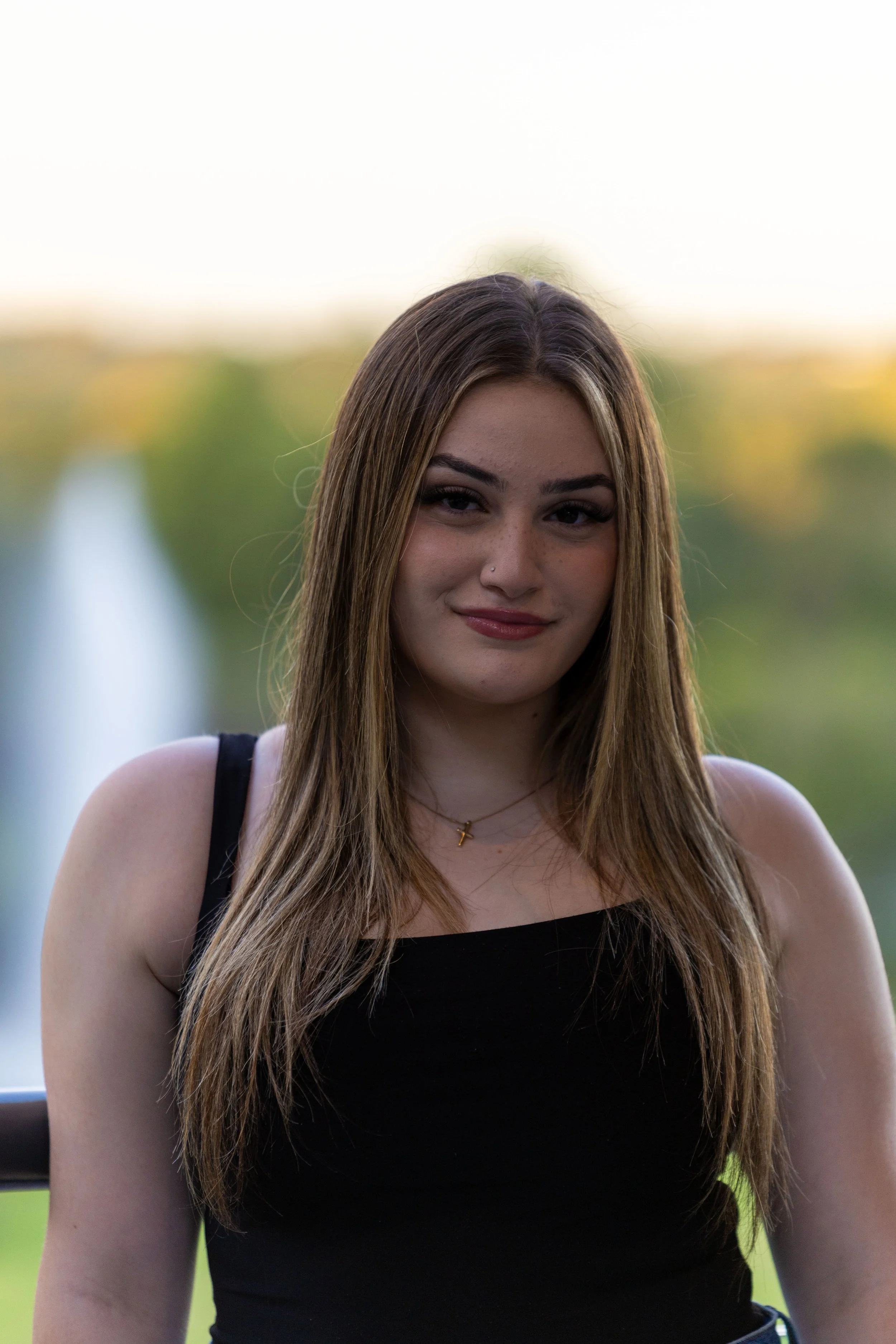 Young woman with long brown hair, wearing a black sleeveless top, standing outdoors with a blurred background of greenery and water fountain.