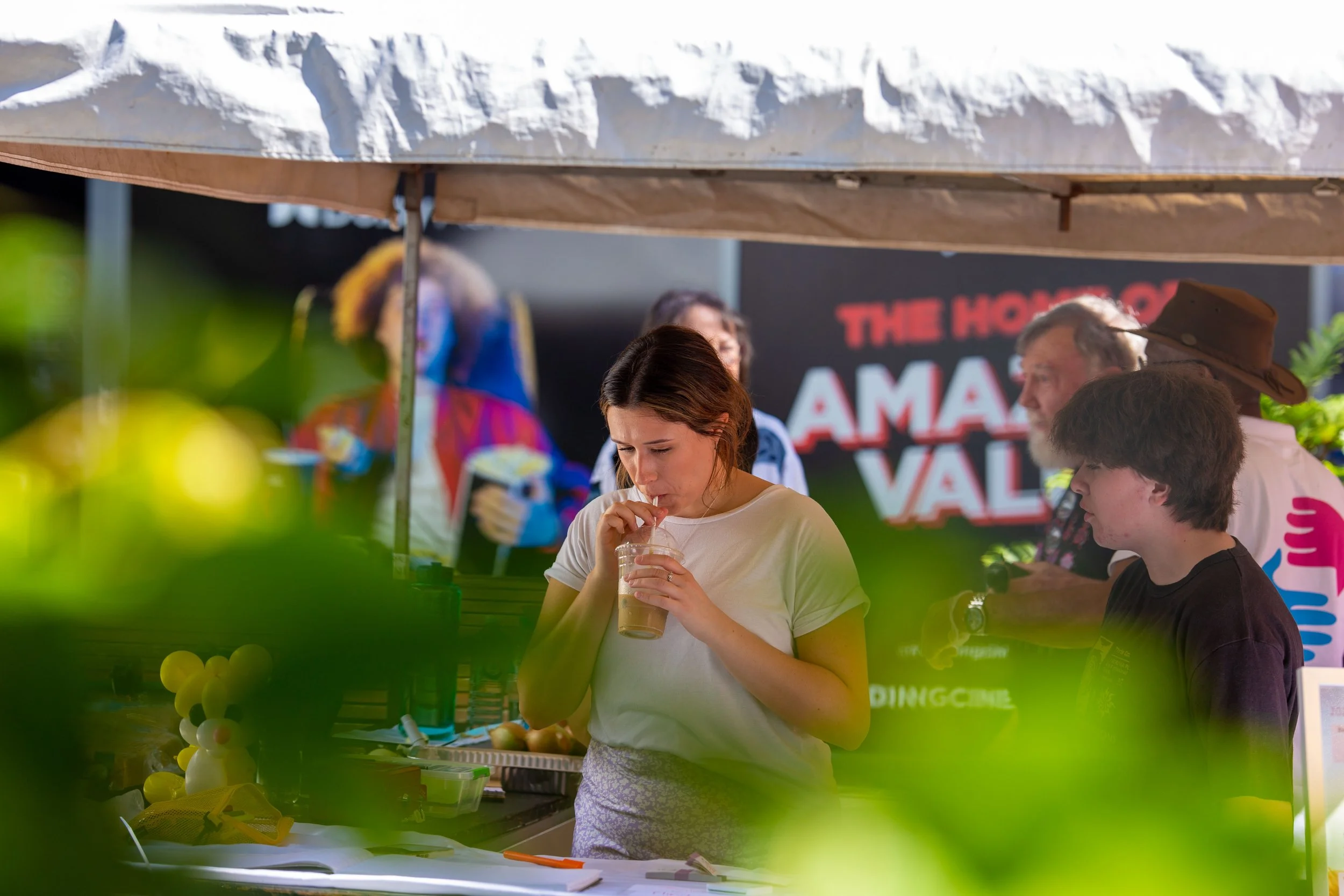 A young woman with brown hair drinking from a clear plastic cup with a straw at an outdoor event or festival, under a canopy tent, with a background of people and a sign that reads "The Homestead Amazing Valley".