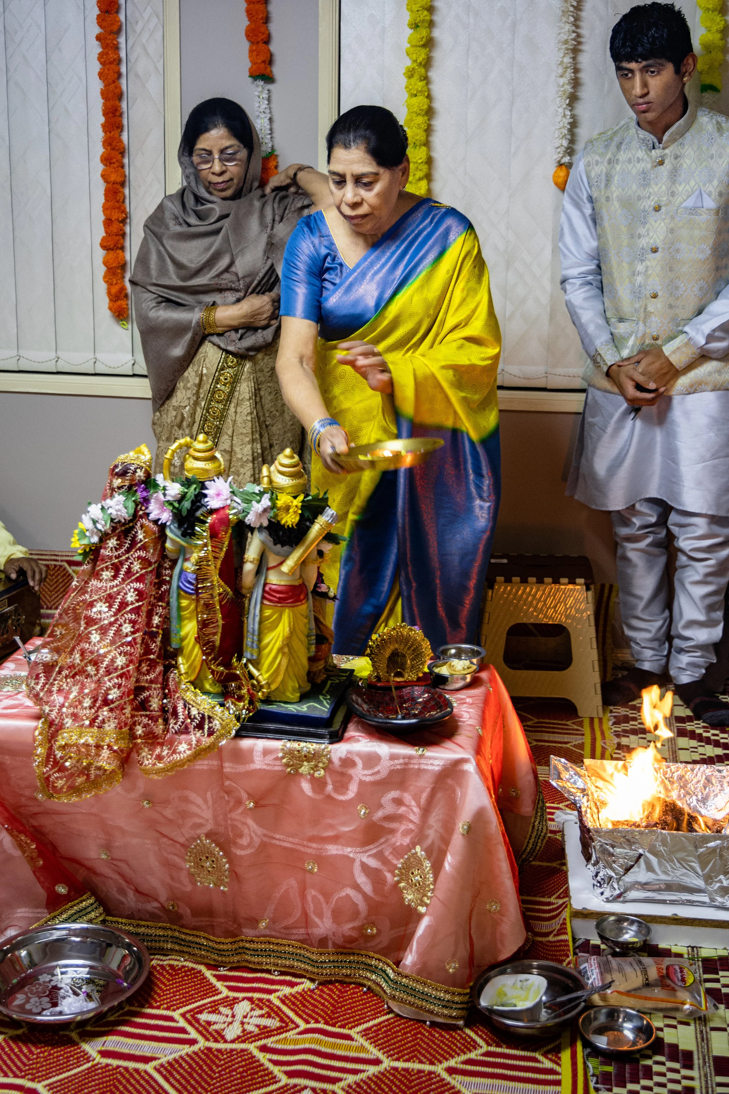 Three people participate in a Hindu religious ceremony indoors. The woman in the center, dressed in a blue and yellow sari, is performing a ritual with a brass lamp. Two other women and a young man observe, with an altar in front that includes decora