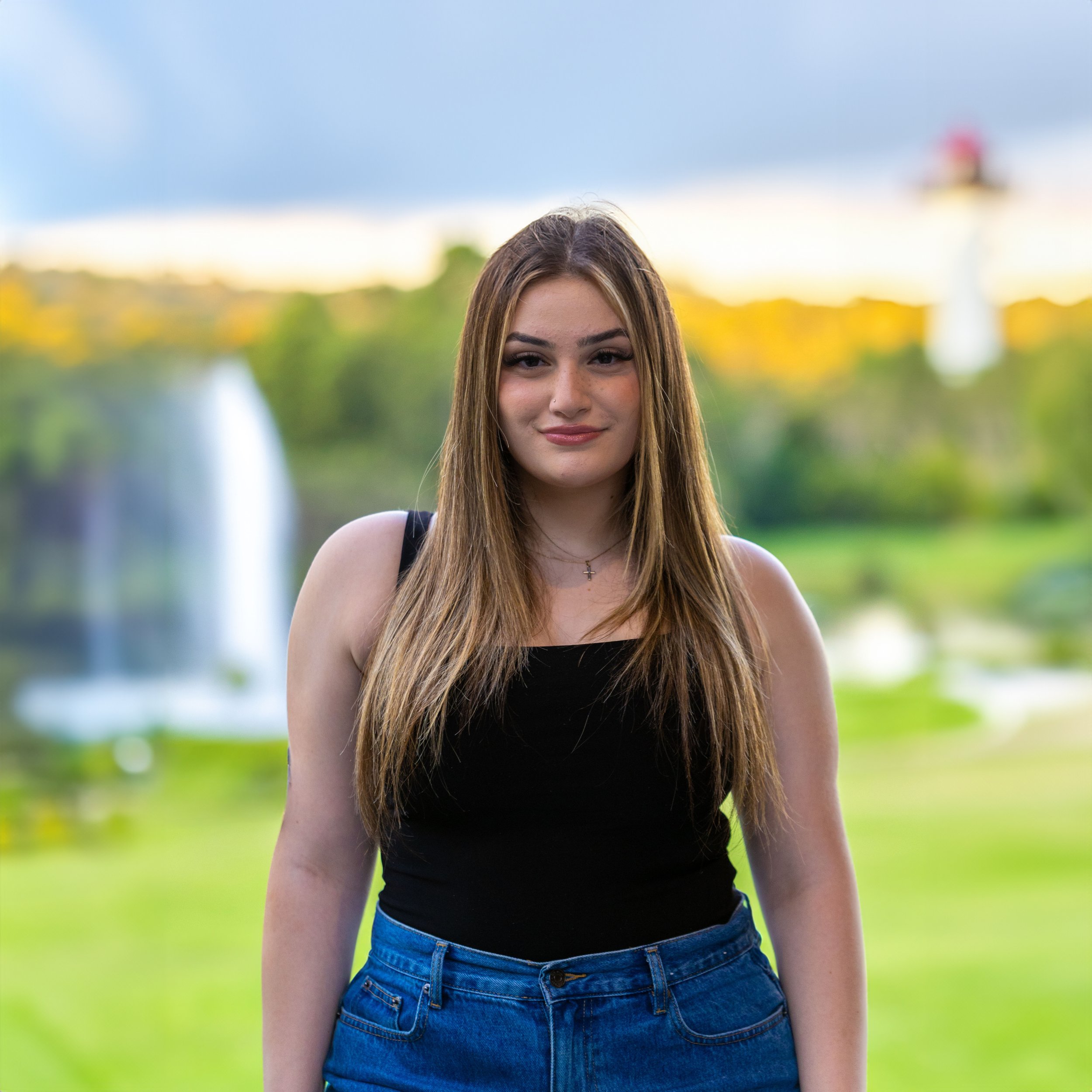 A young woman with long brown hair standing outdoors in front of a blurred background of a park, water fountain, green trees, and a lighthouse. She is wearing a black tank top and blue jeans, and has a gentle smile.