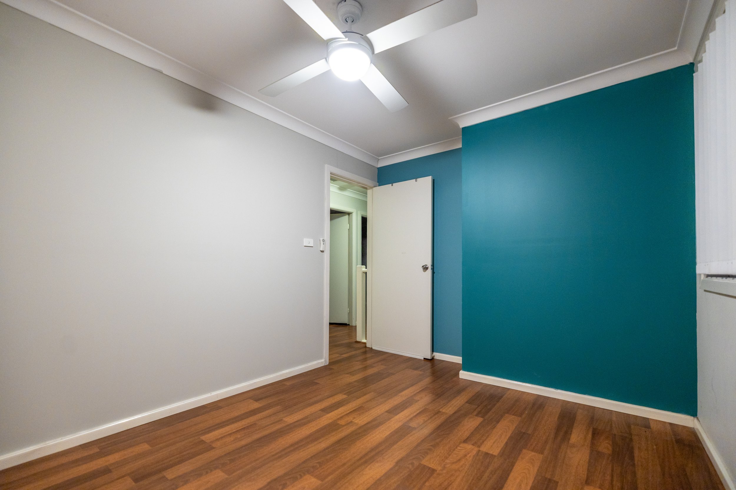 Empty room with white and teal walls, wooden floor, ceiling fan with light, and an open door showing a hallway.