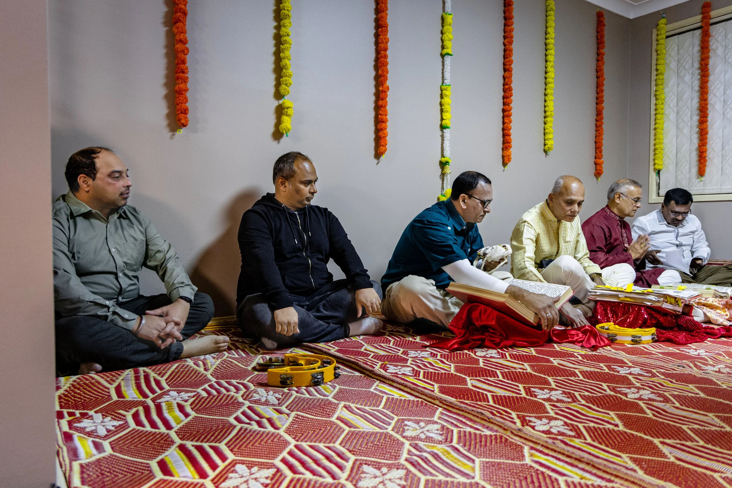 Six men sitting on the floor during a traditional ceremony, with marigold and other flower garlands hanging on the wall behind them. Some men are reading from books or scriptures, and there are musical instruments like tambourines placed on the floor
