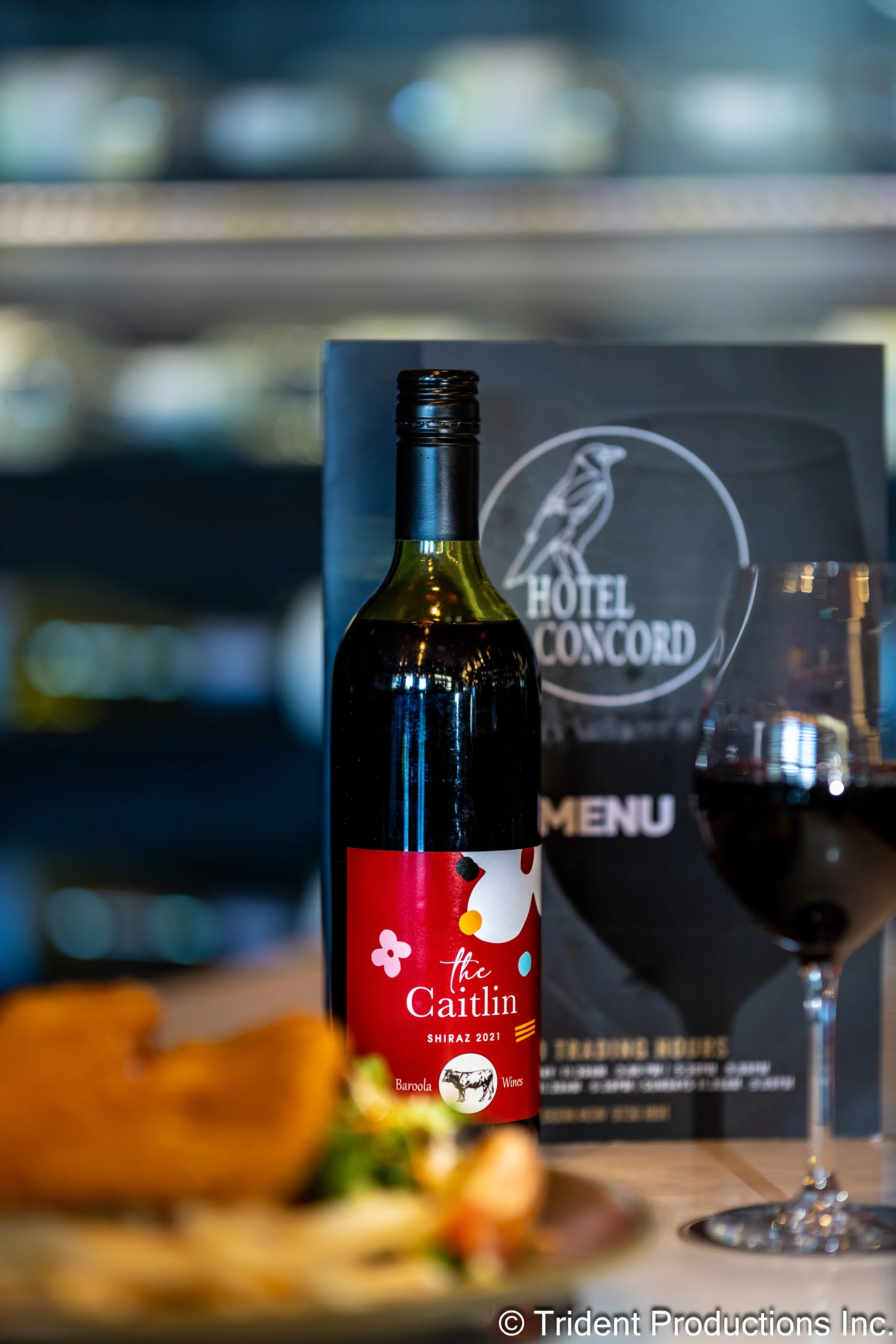 A bottle of red wine labeled 'The Caitlin', a glass of red wine, a menu from Hotel Concord, and a plate of food in the foreground.