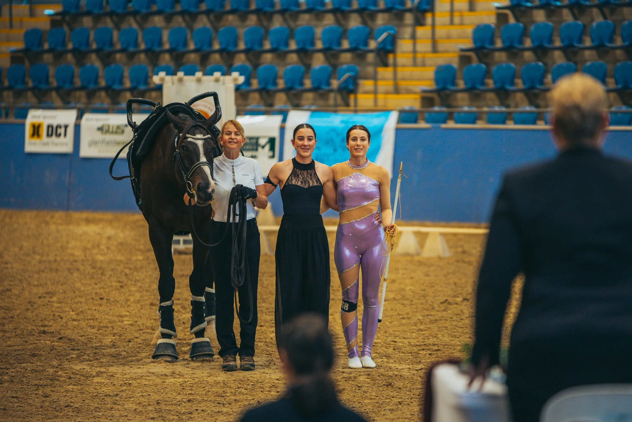 Three women and a horse standing in an indoor arena. The women are smiling, with the woman on the left holding the horse's reins. The woman on the right wears a purple, shiny athletic outfit, and the middle woman is in a black dress. There are specta