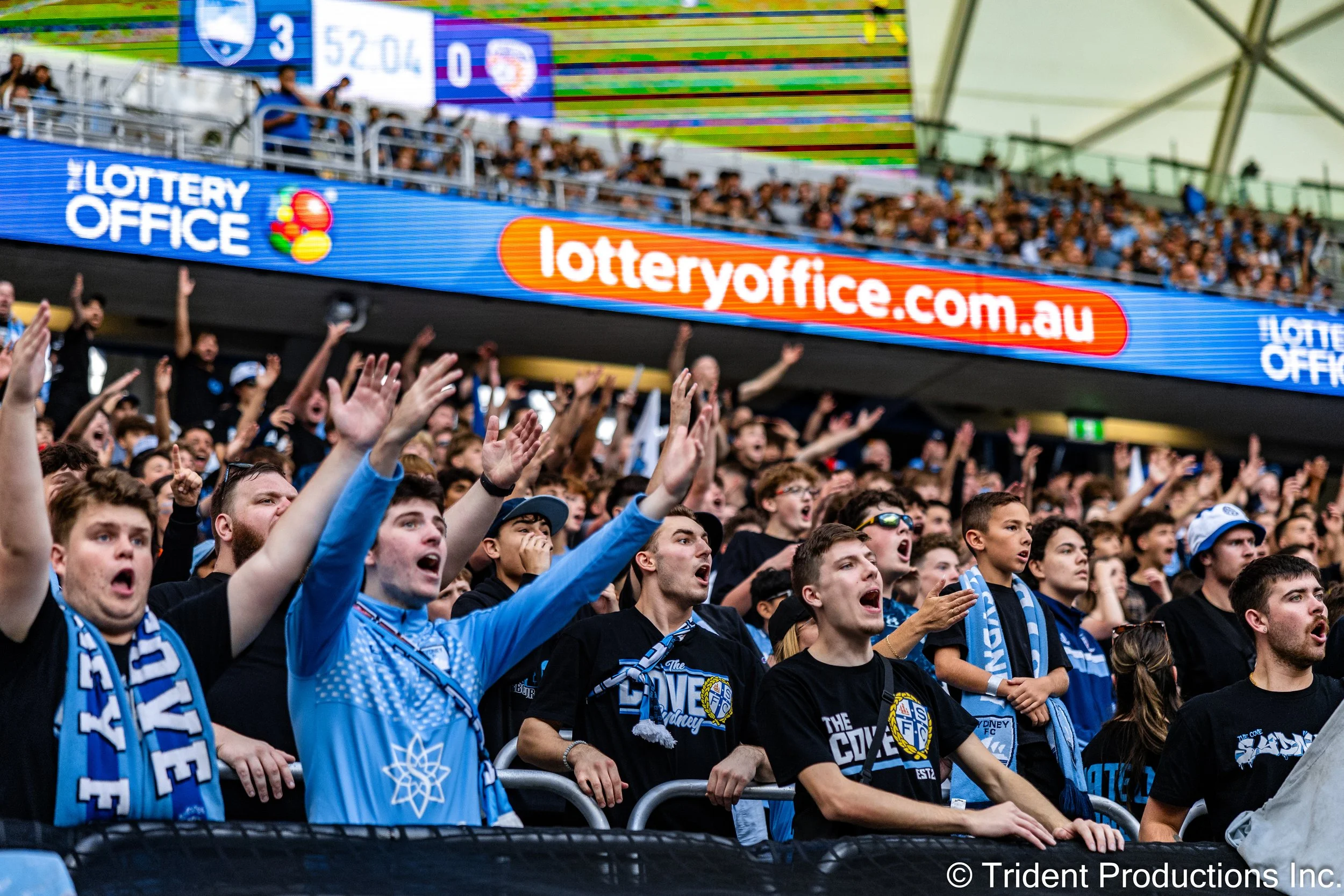 Crowd of sports fans wearing Sydney FC jerseys and scarves, cheering at a soccer match in a stadium with a digital advertising banner displaying 'lotteryoffice.com.au'.