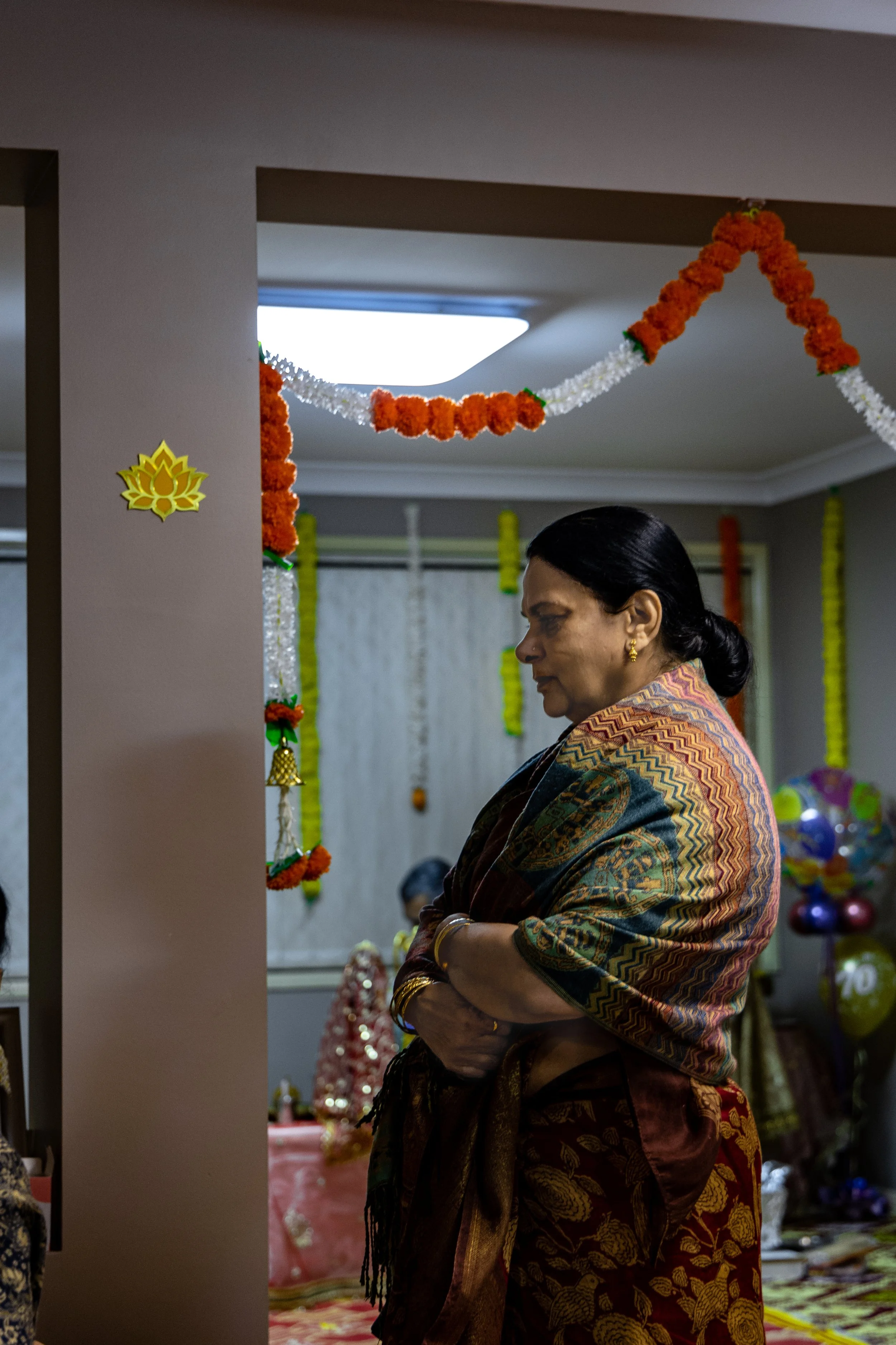 Indian woman wearing traditional saree and jewelry, standing with folded arms inside a decorated room for a celebration.