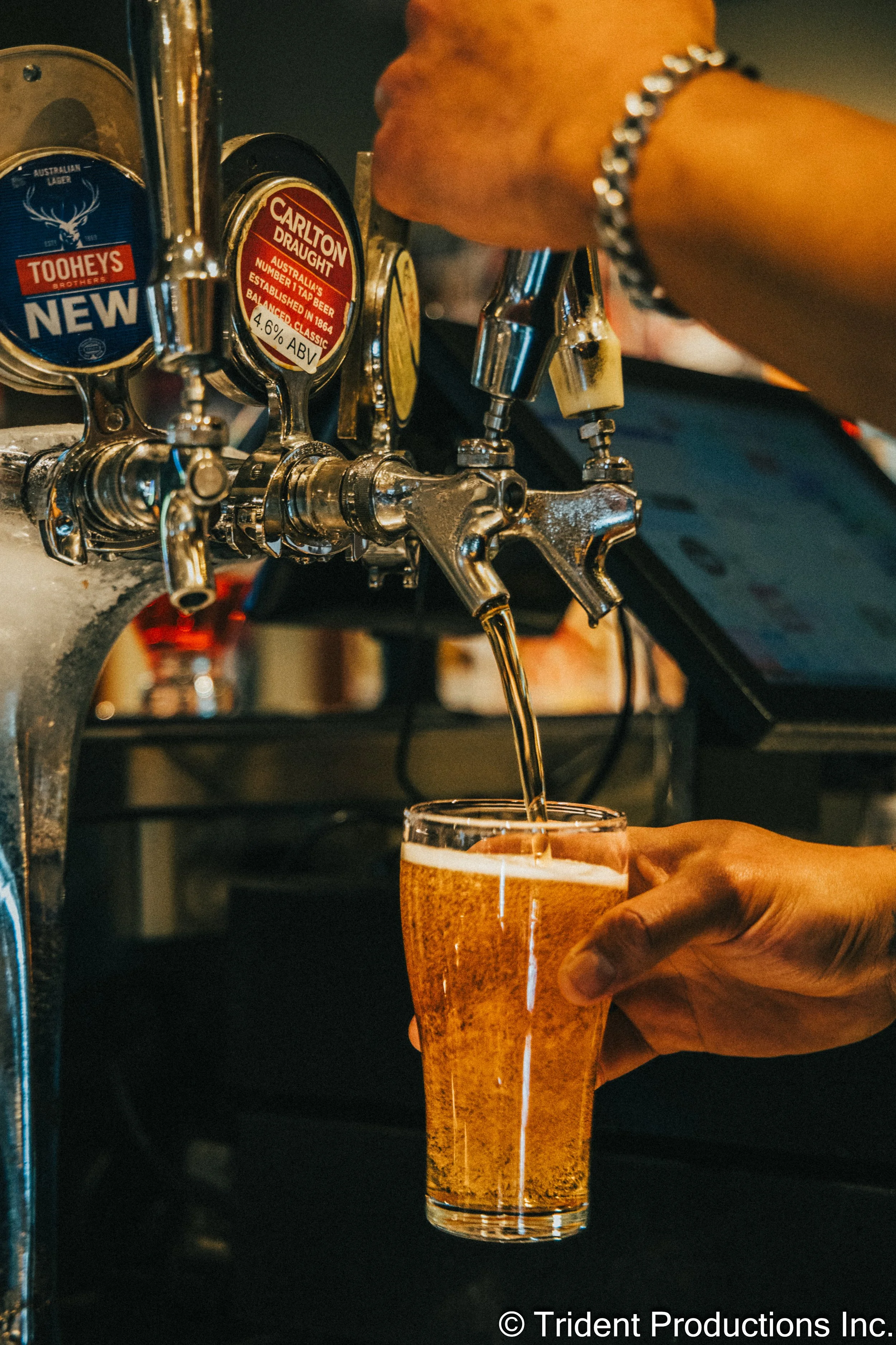 A person is pouring beer from a tap into a glass at a bar or pub. The image shows multiple beer taps, and the person's hand is holding the glass, filling it with beer.