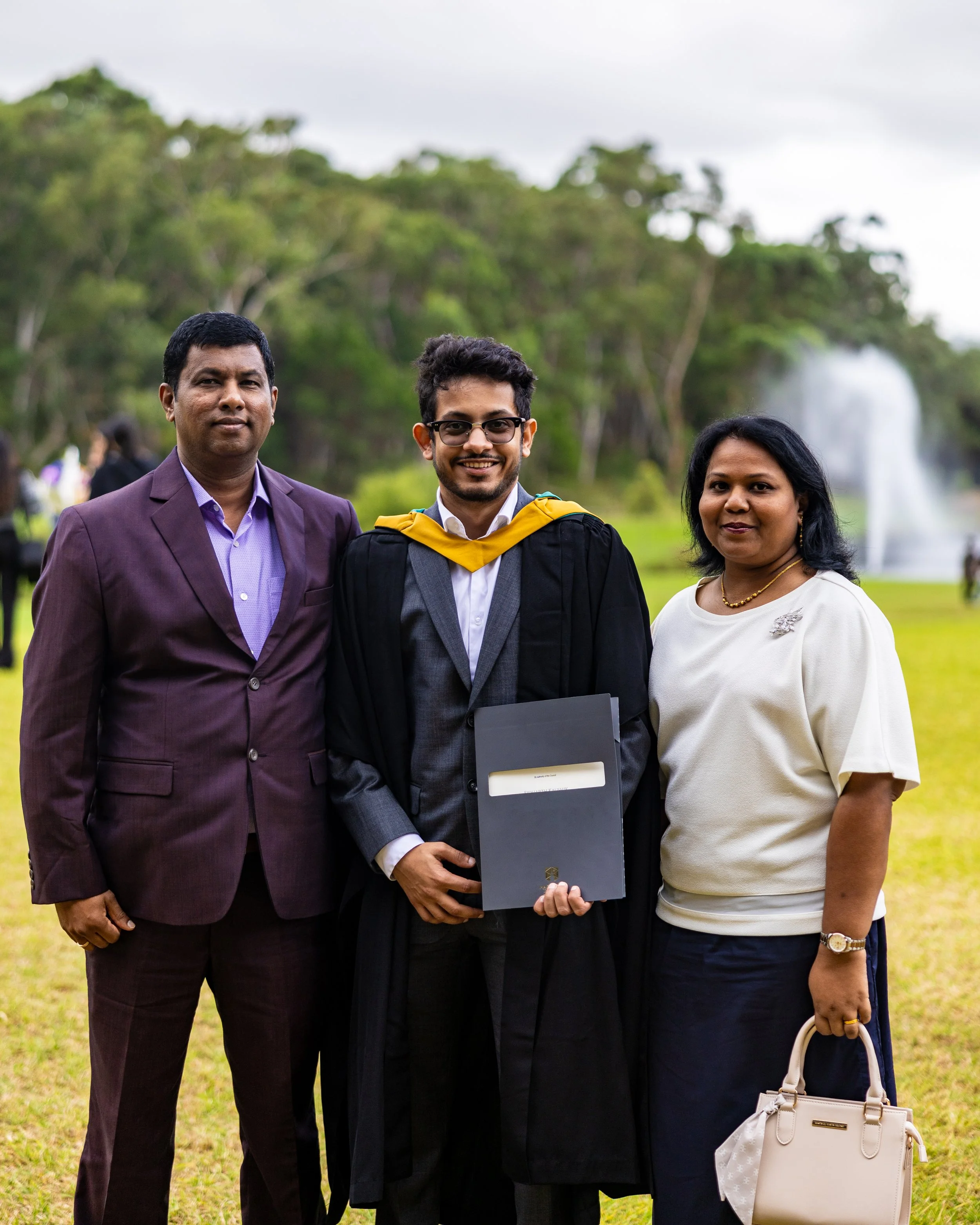 A young man in graduation gown and cap with a yellow sash, holding a diploma, standing between a man in a purple suit and a woman in white blouse at an outdoor graduation ceremony.