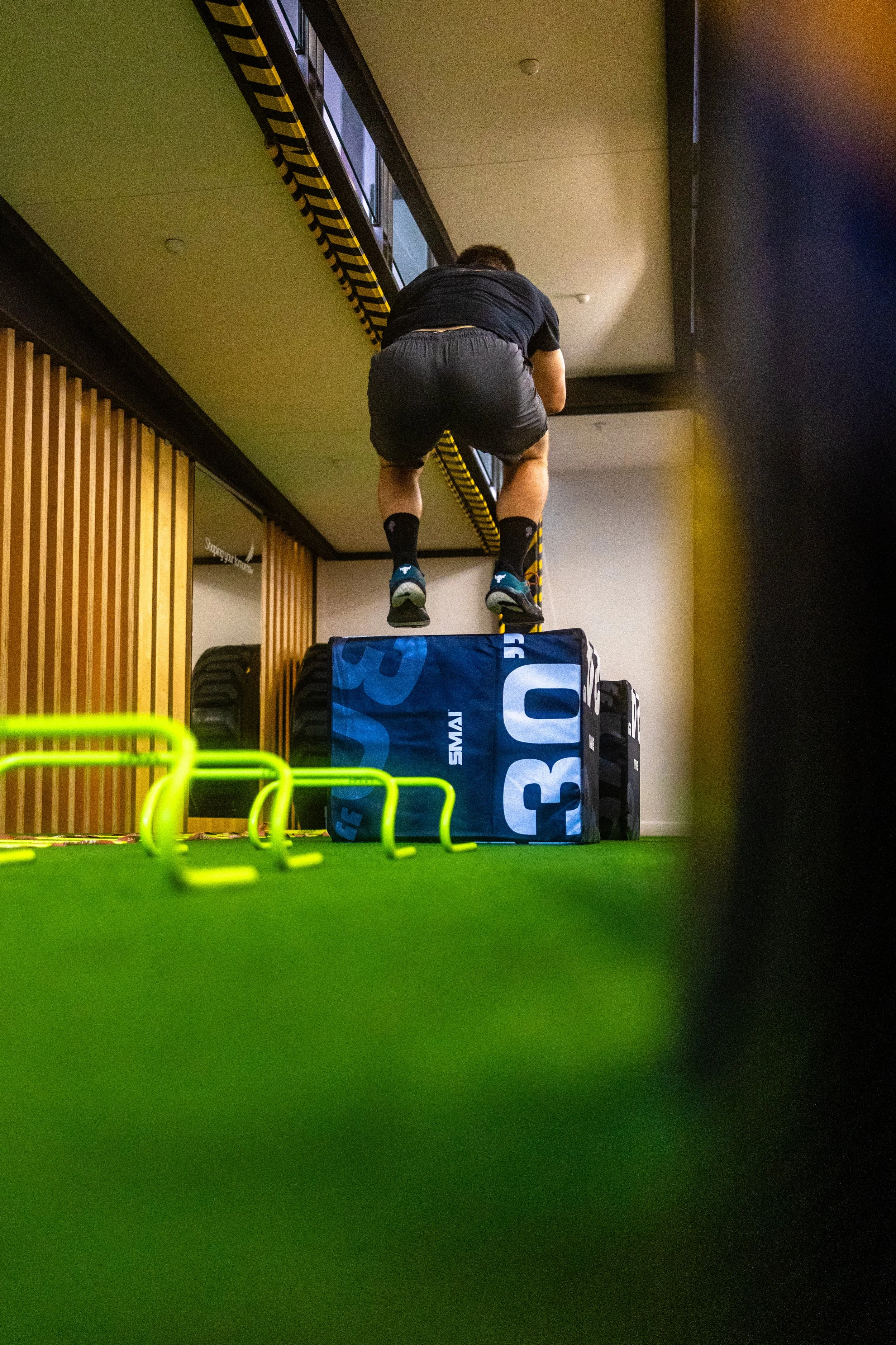 A person performing a workout in an indoor gym, climbing over a blue exercise box, with green hurdles on the green turf in the foreground.