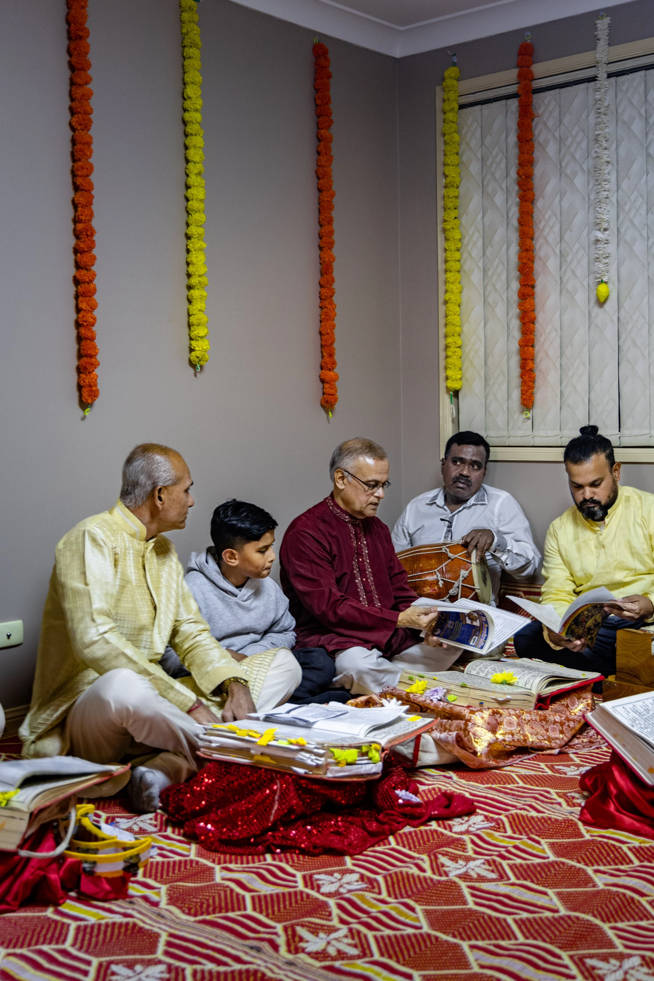 Group of six men and one boy sitting on the floor during a Hindu religious ceremony, reading scriptures, with garlands hanging on the wall and a decorative red and gold cloth covering the floor.