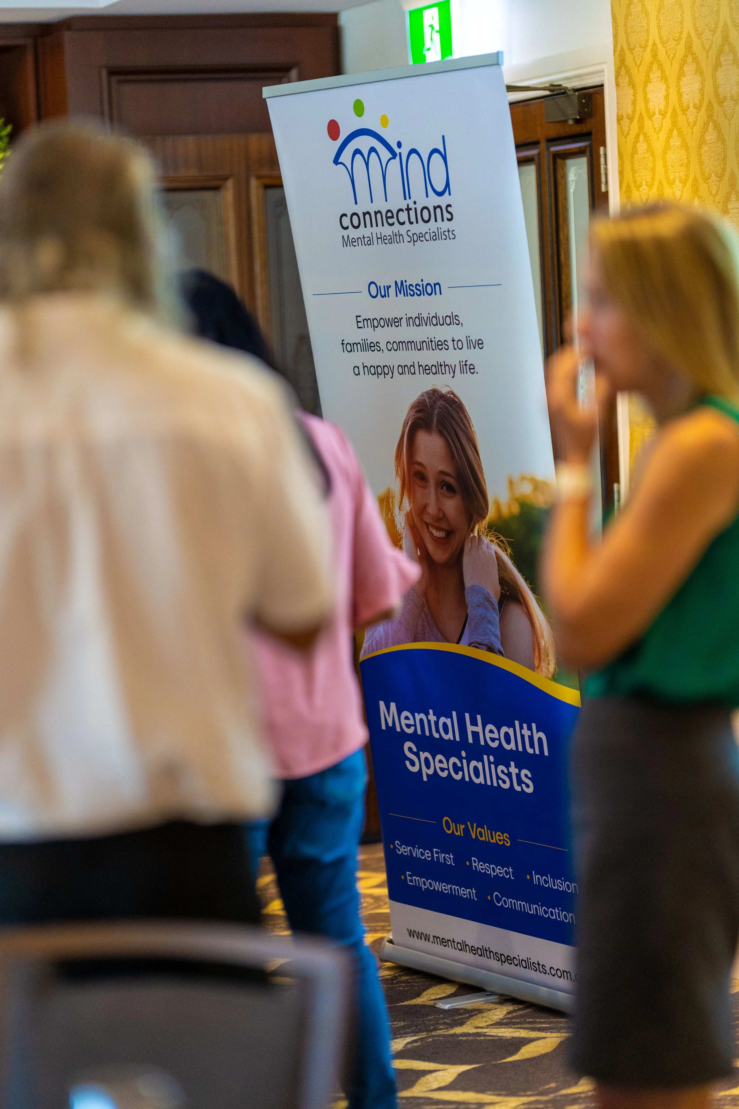 A group of people gathered around a banner for a mental health organization called 'Mind Connections'. The banner displays the organization's mission statement and values, focusing on empowering individuals and communities for a healthy life. The set