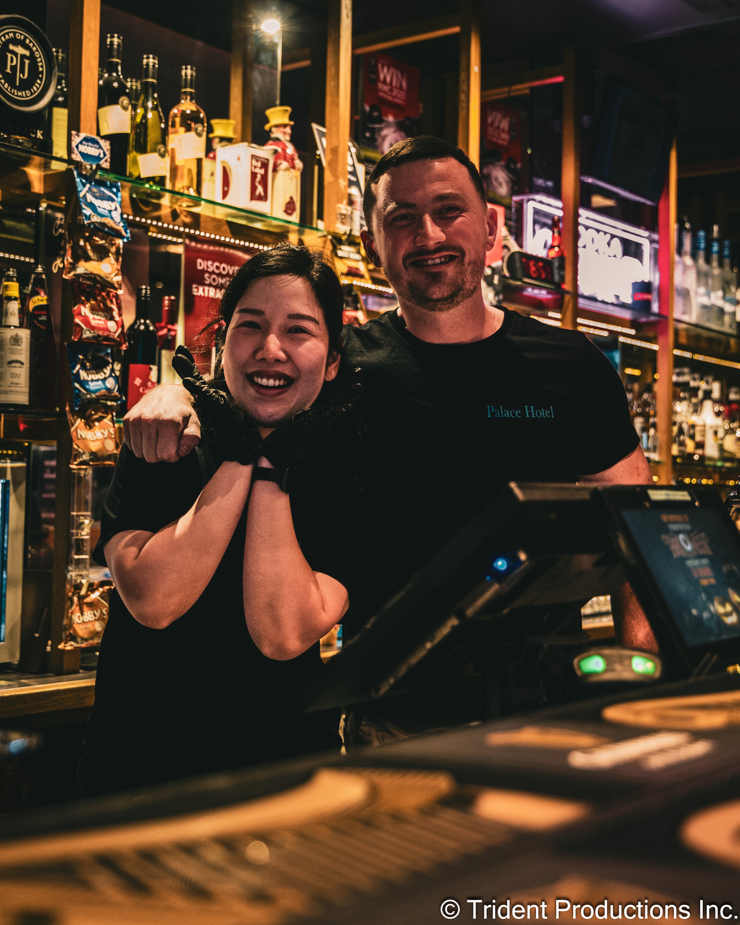 Two bartenders, a woman and a man, smiling behind a bar at a pub, with liquor bottles and snacks on shelves in the background.