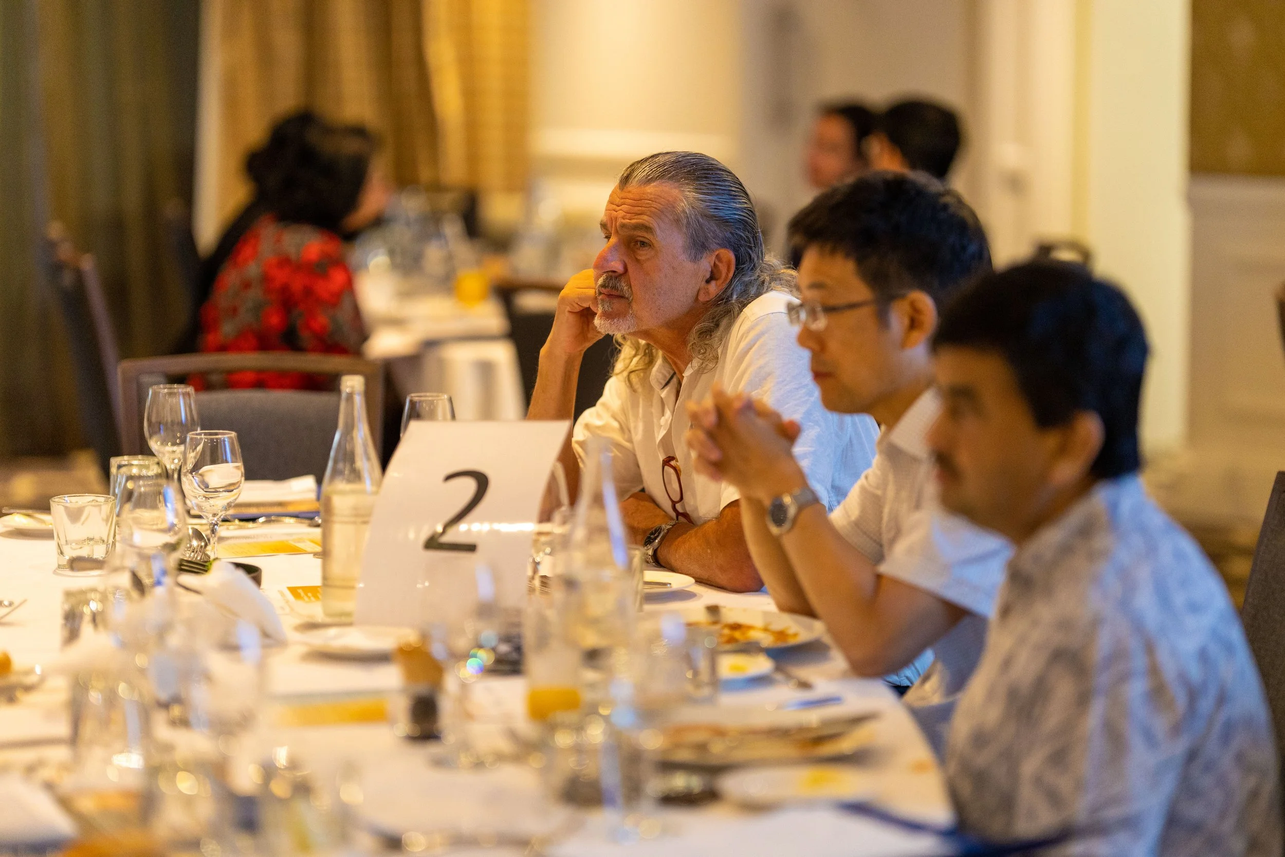 People sitting at a banquet table paying attention, with a middle-aged man with gray hair and a beard in focus.