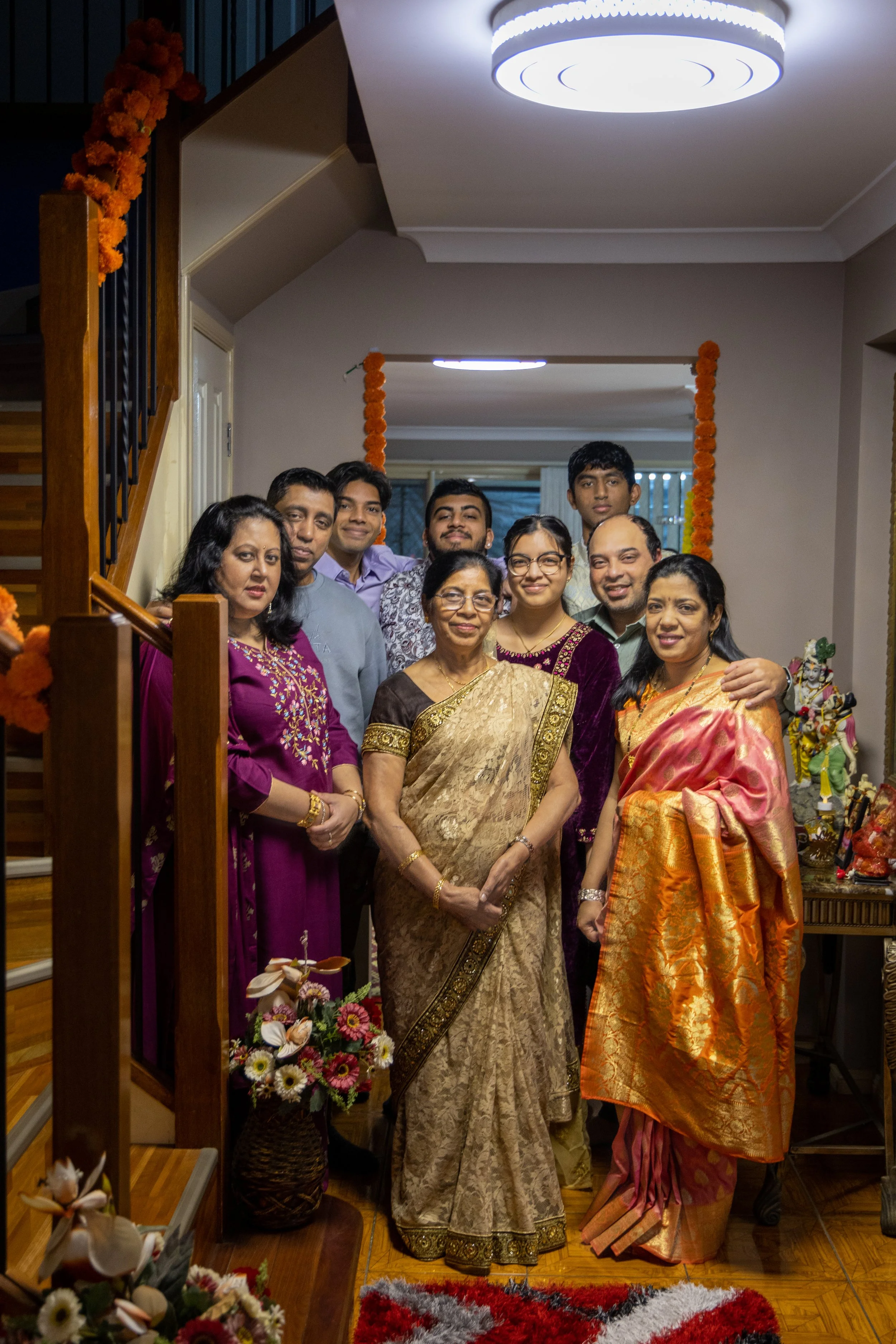 Family gathered together in a living room decorated for a celebration, with a floral arrangement and Hindu deity statues visible.