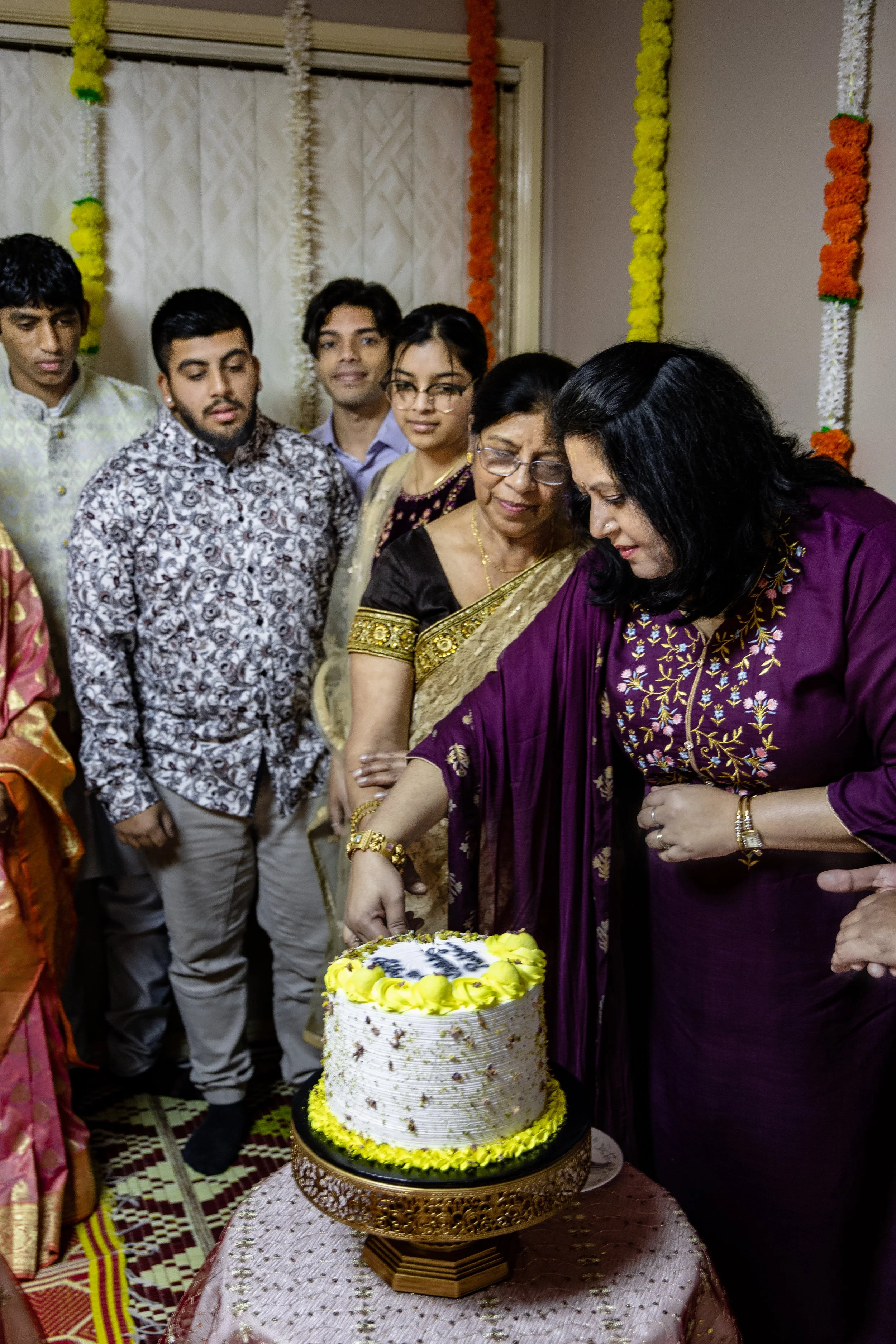 A group of people gathered around a decorated cake during a celebration, with women cutting the cake and men standing behind them, inside a room decorated with colorful flowers and streamers.