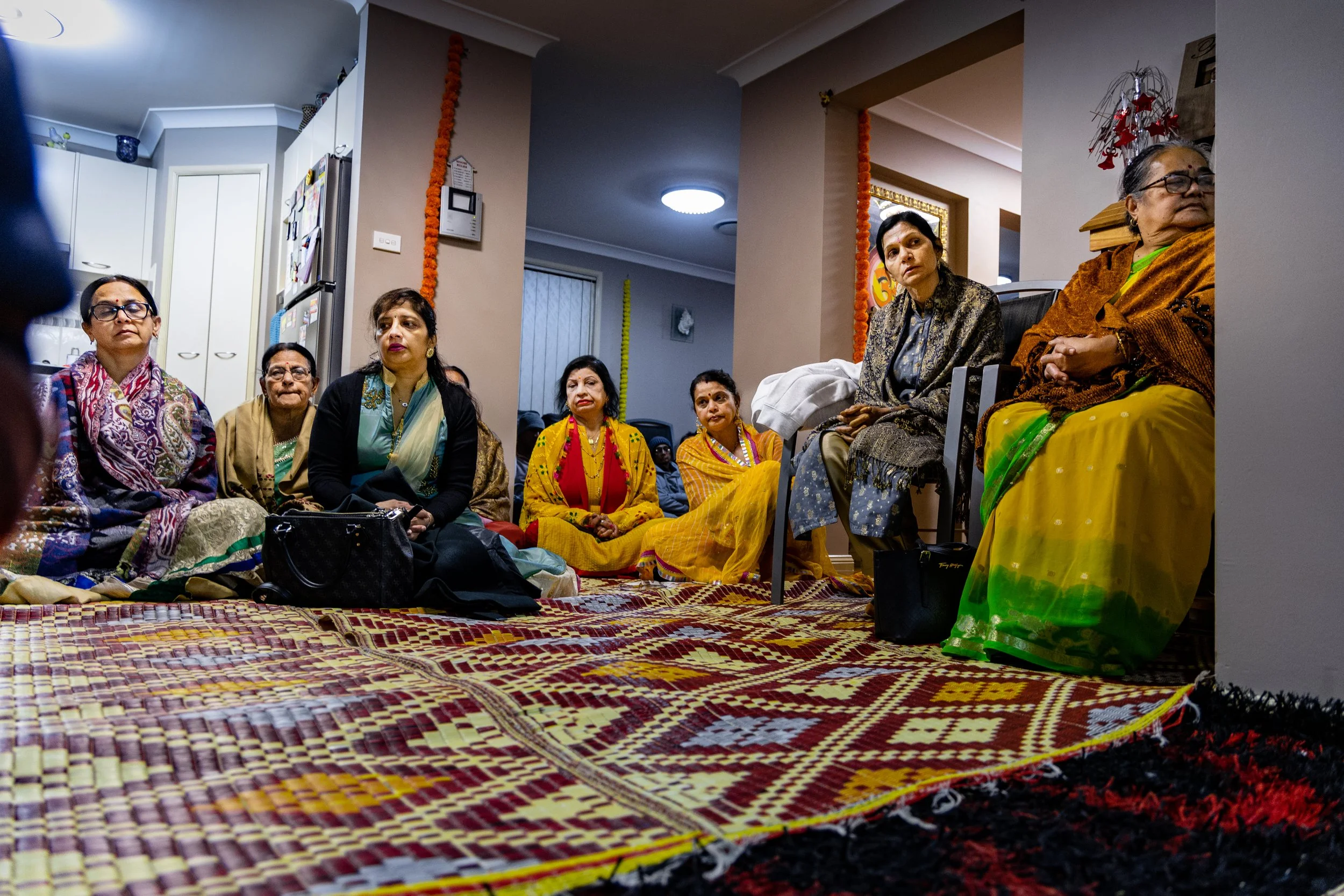 A group of women sitting on the floor and on chairs inside a room, possibly attending a cultural or social gathering. They are dressed in colorful traditional Indian attire, including sarees and shawls.