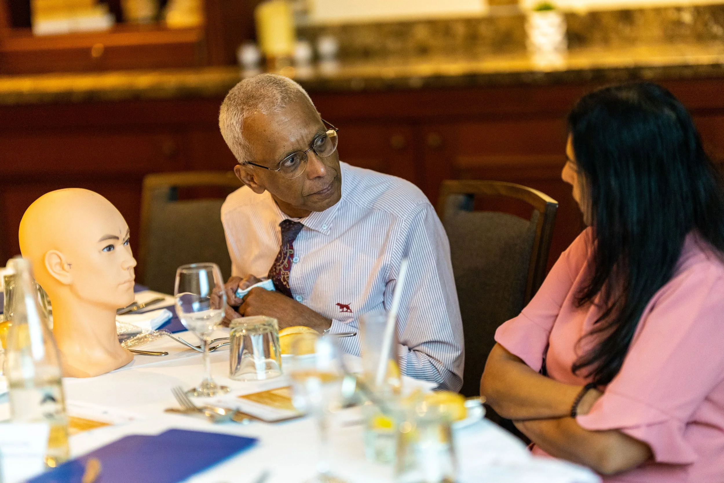 An elderly man with glasses and a woman in conversation at a table set for a meal, with a mannequin head on the table, in a restaurant or banquet hall.