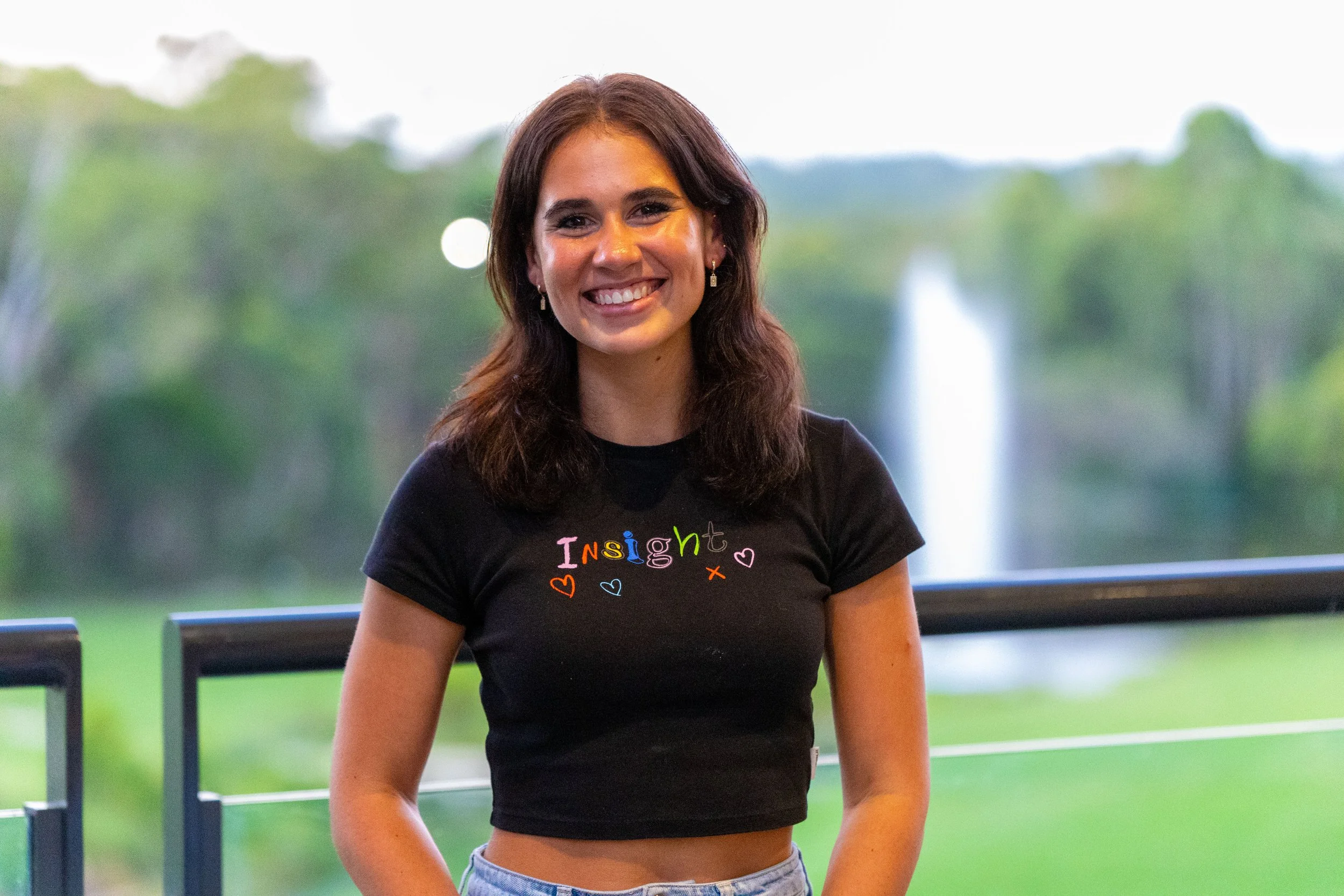 A young woman with shoulder-length brown hair and earrings, smiling, wearing a black t-shirt with colorful text and hearts, standing outdoors with greenery and a fountain in the background.
