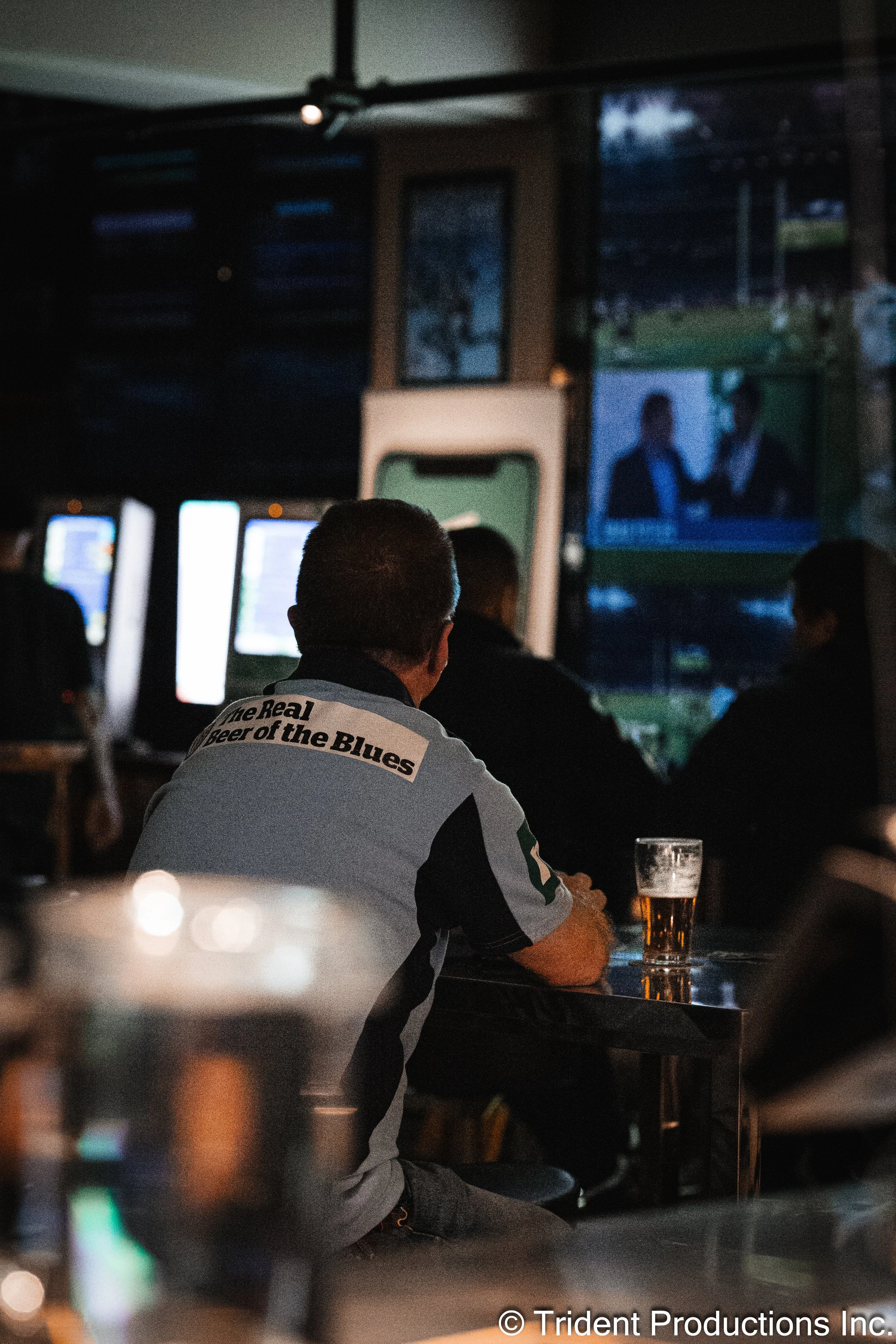 A man sitting at a bar watching a sports game on television, with a pint of beer in front of him. The bar appears dimly lit, and other patrons are also watching the game.