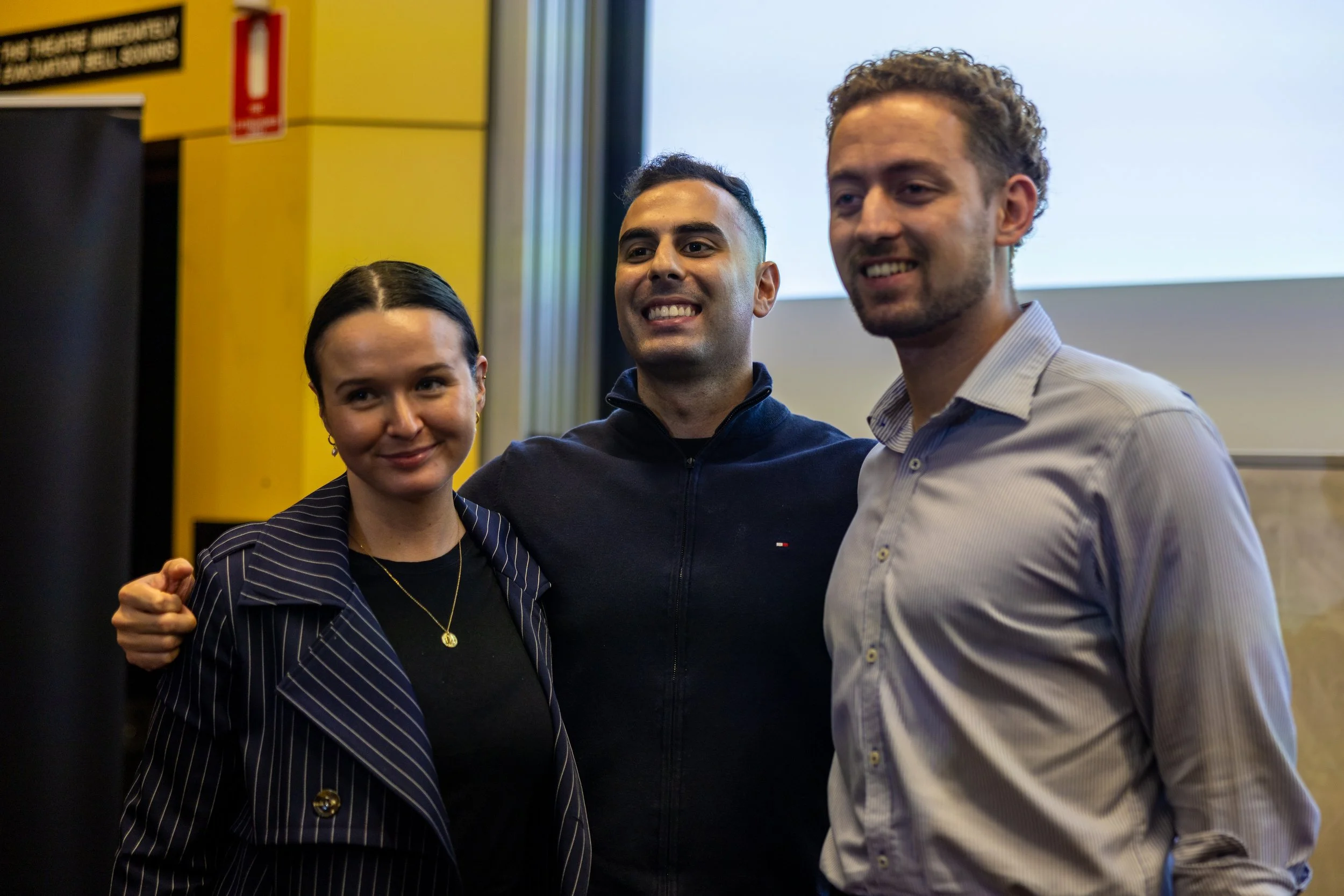 Three people standing close together, smiling at a photo opportunity, in a room with a yellow wall and a projection screen.