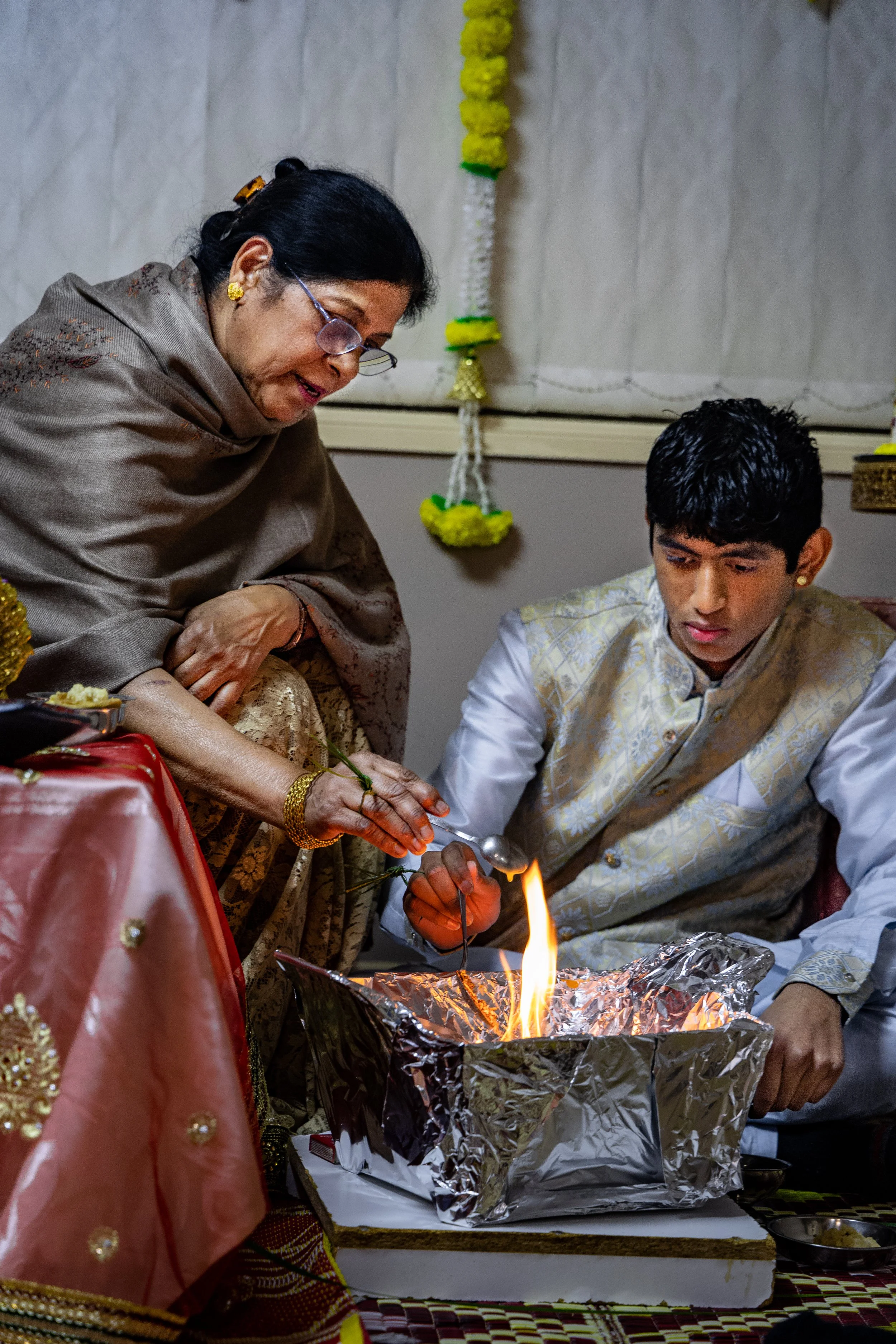 A woman and a young man participating in a traditional Indian ceremony, with the woman holding a spoon with fire over a silver container, as part of a ritual.