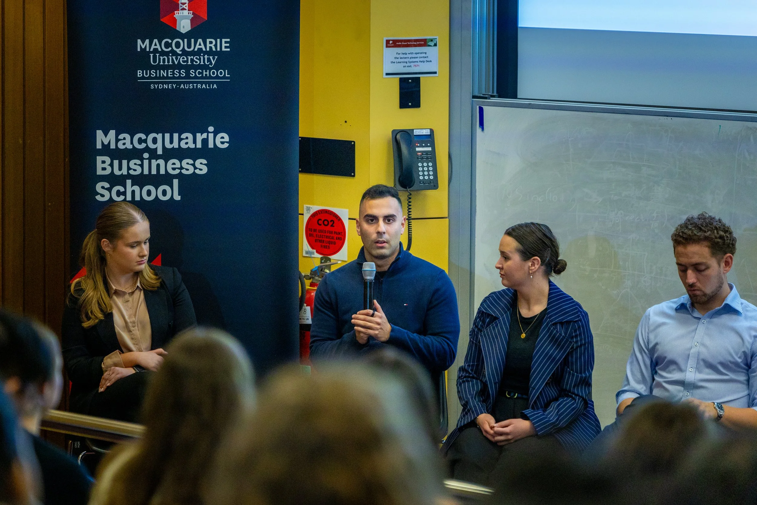 A panel discussion at Macquarie University Business School in Sydney, Australia, with four people seated or standing near a banner and a chalkboard.