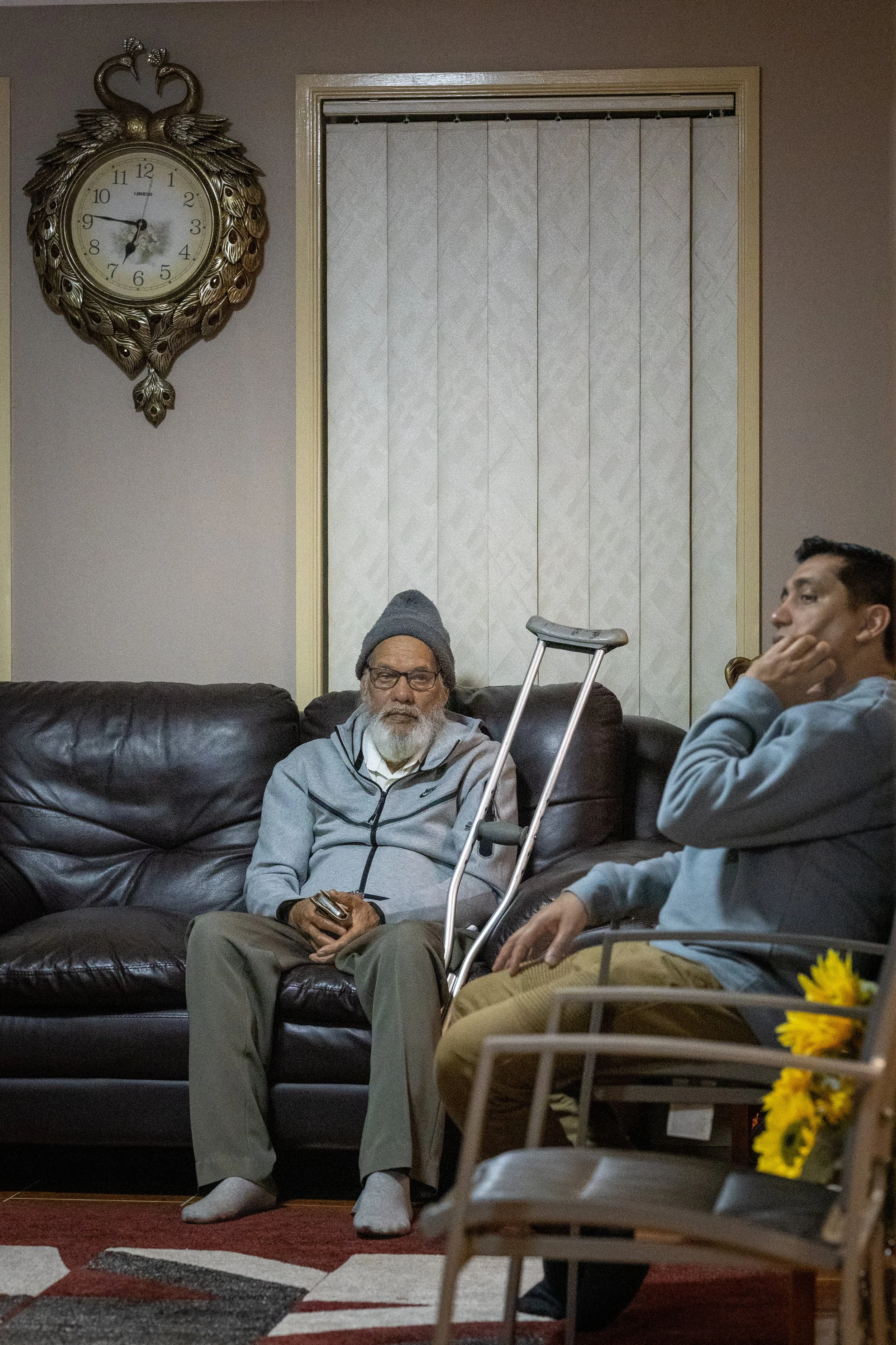 Two men sitting in a living room with a clock on the wall, one man is elderly with a gray beard, wearing glasses, a gray beanie, jacket, and sitting on a leather couch holding a remote; the other man is younger, resting his face on his hand, sitting 