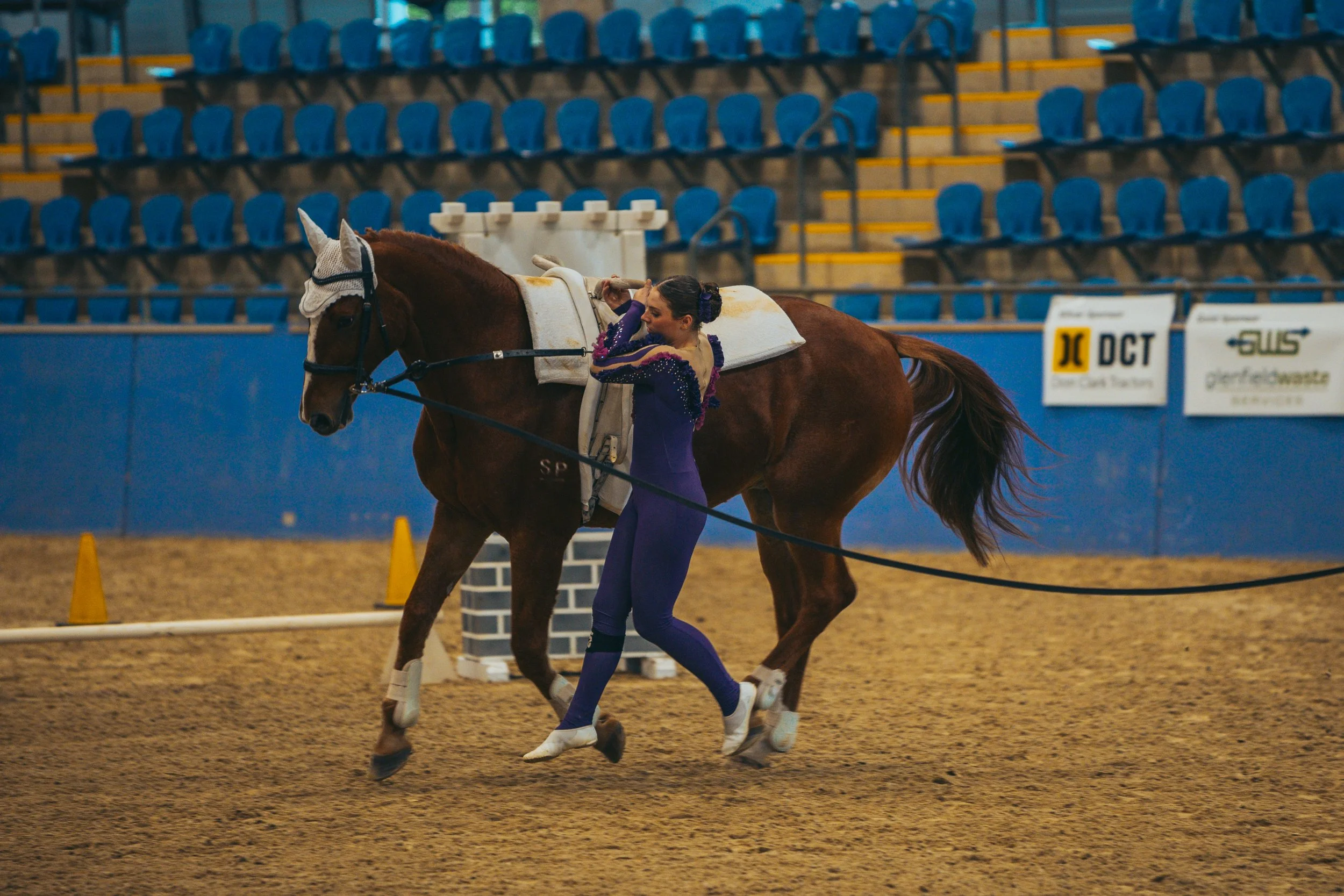 A female equestrian performer adjusting her costume next to a chestnut horse inside an indoor arena with blue seats and advertising banners.