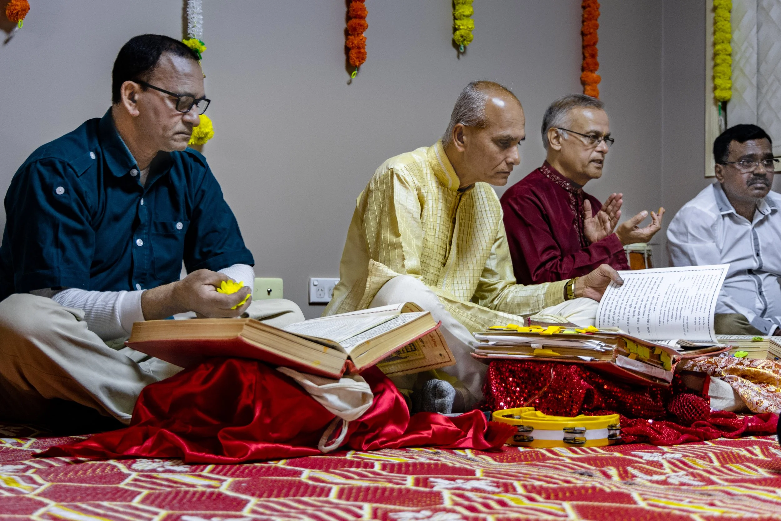 Four men sitting cross-legged on a patterned red and gold floor, engaging in a religious or ceremonial activity. They are dressed in traditional Indian attire, with flowers and books or scriptures in front of them. The background is decorated with ha