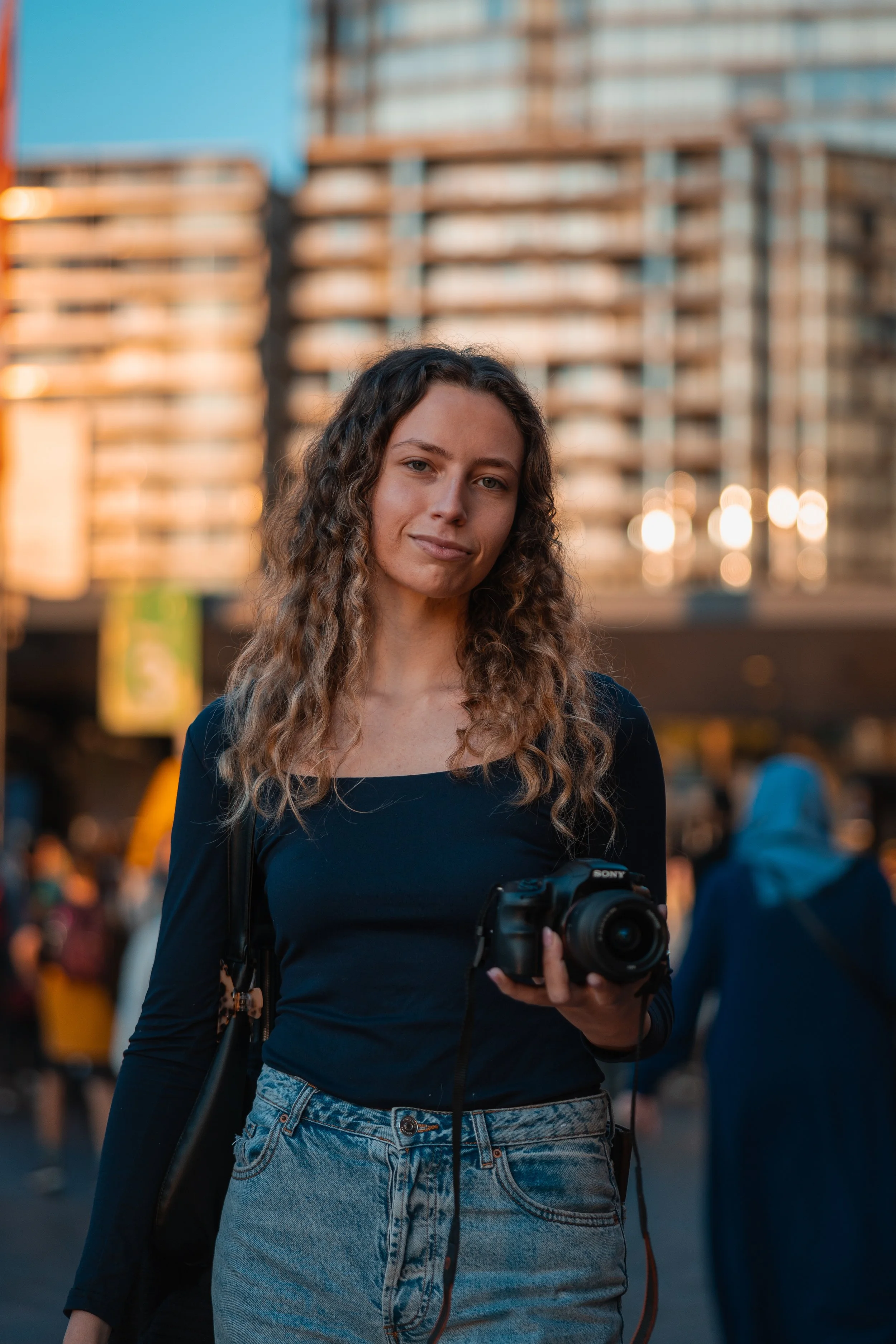 A young woman with curly hair holding a Sony camera in an urban setting during sunset, with tall buildings and people in the background.