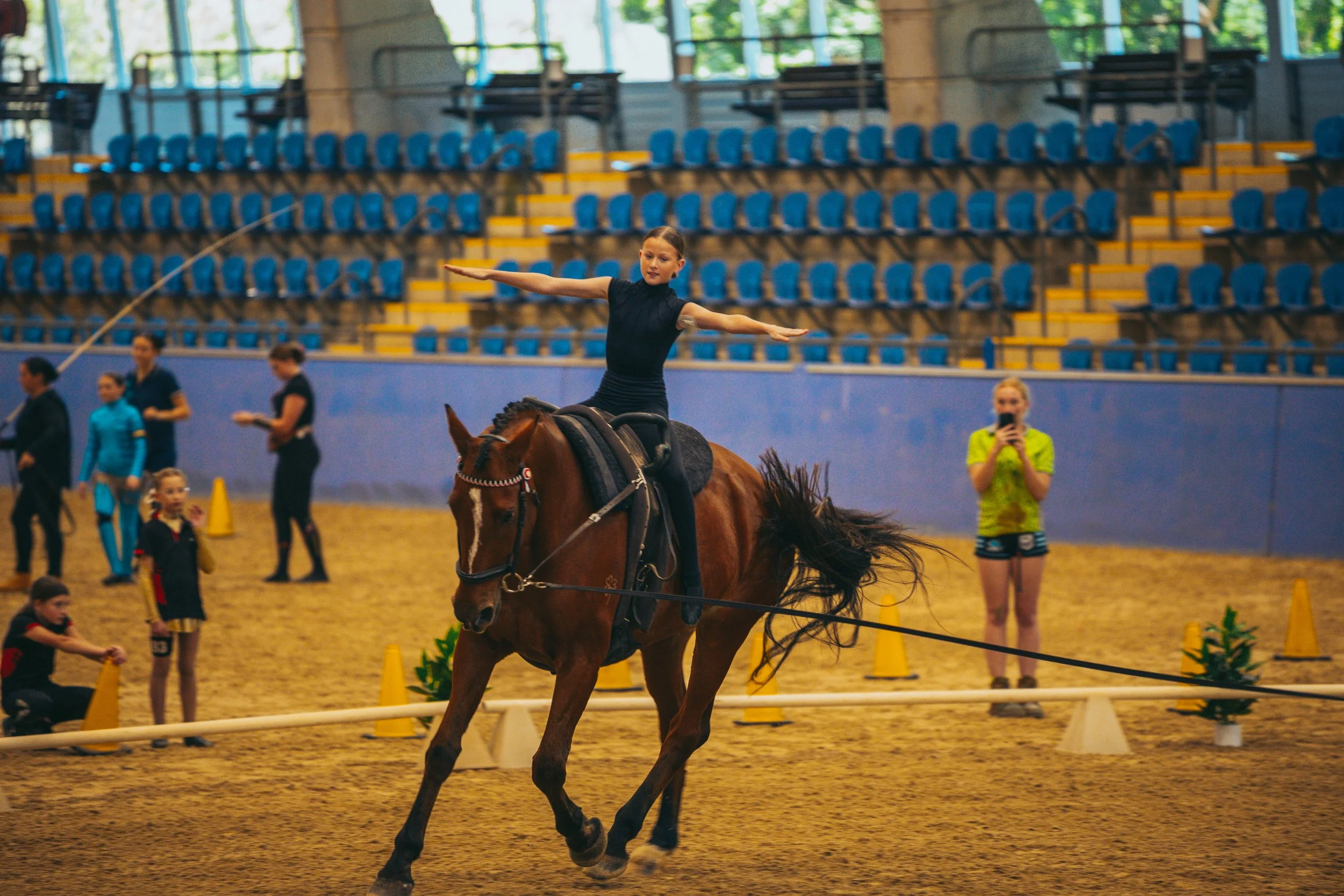 A young girl performing a balancing routine on a galloping horse inside an indoor arena, with spectators and officials in the background.