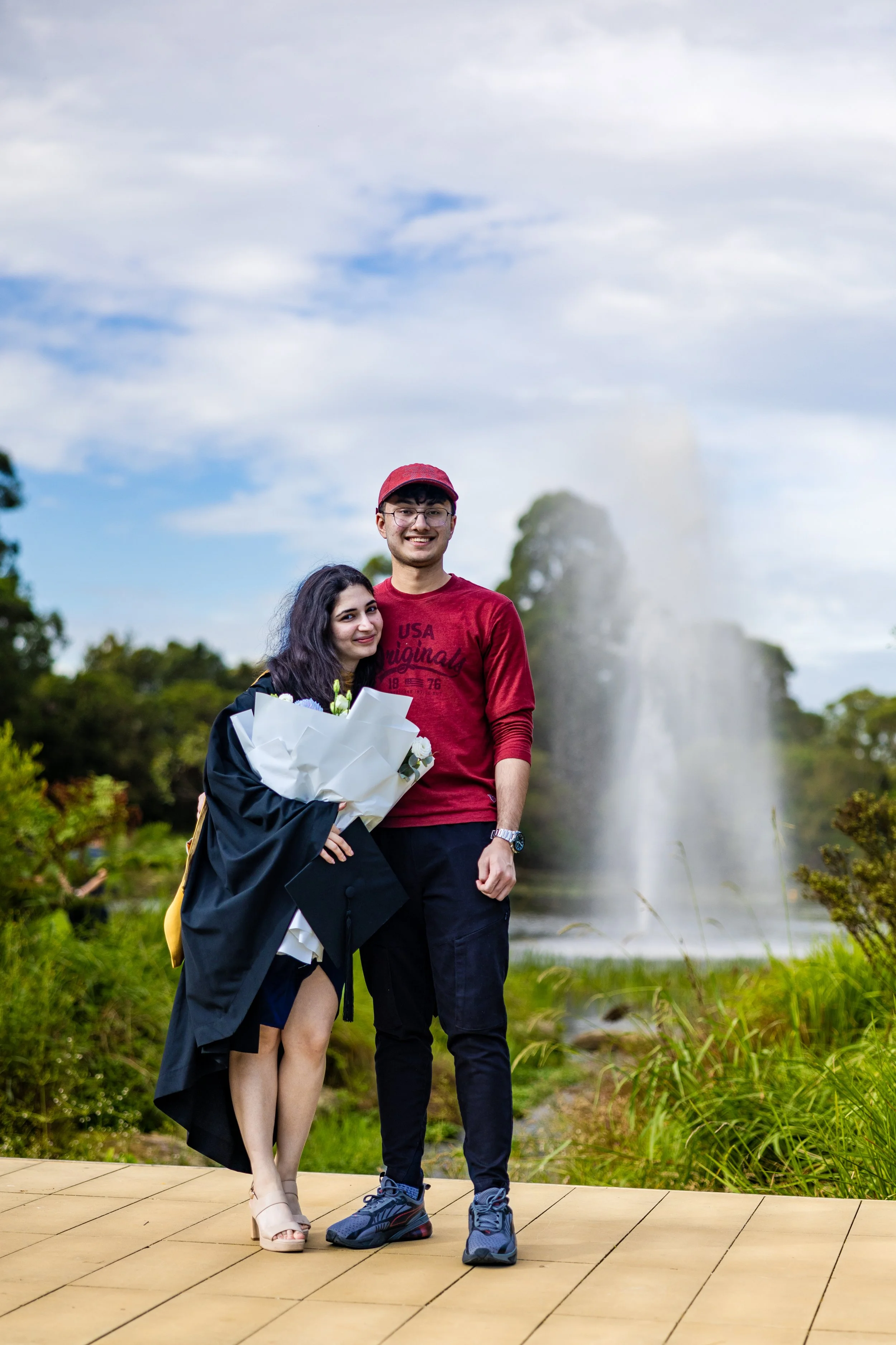 A young woman in a graduation gown and heels holding a bouquet of flowers, and a young man in casual attire, standing together outdoors on a wooden platform with a fountain and greenery in the background.