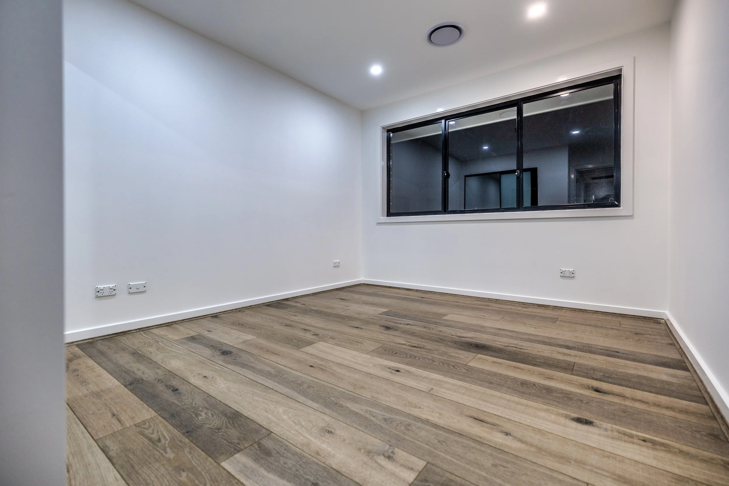 Empty room with white walls, wooden flooring, a large window with a black frame, and some electrical outlets on the lower part of the walls.