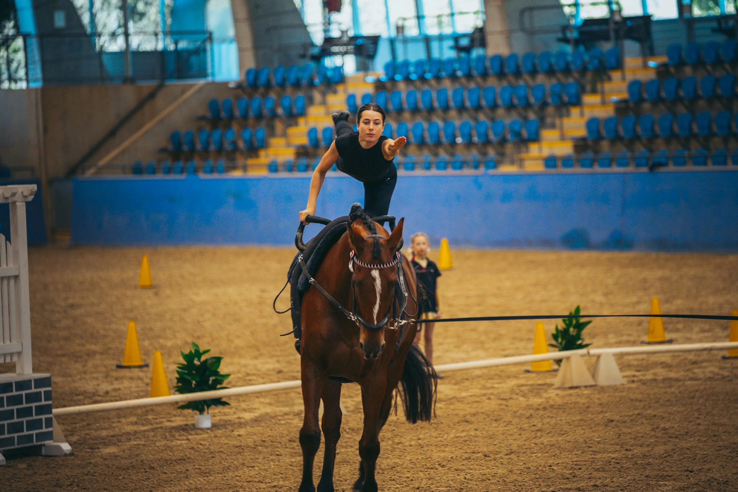 A young woman performs an acrobatic move on a horse inside an indoor equestrian arena, with another girl standing in the background, surrounded by orange cones and green plants.