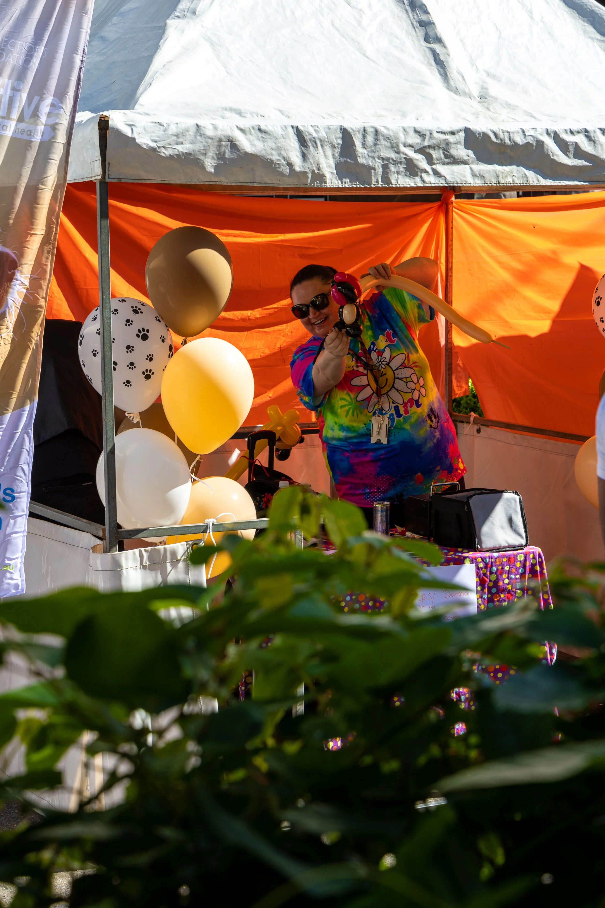 A woman in a colorful tie-dye shirt and sunglasses is standing at a booth decorated with balloons, some paw print patterned, under an orange canopy. She is holding a balloon animal and smiling.