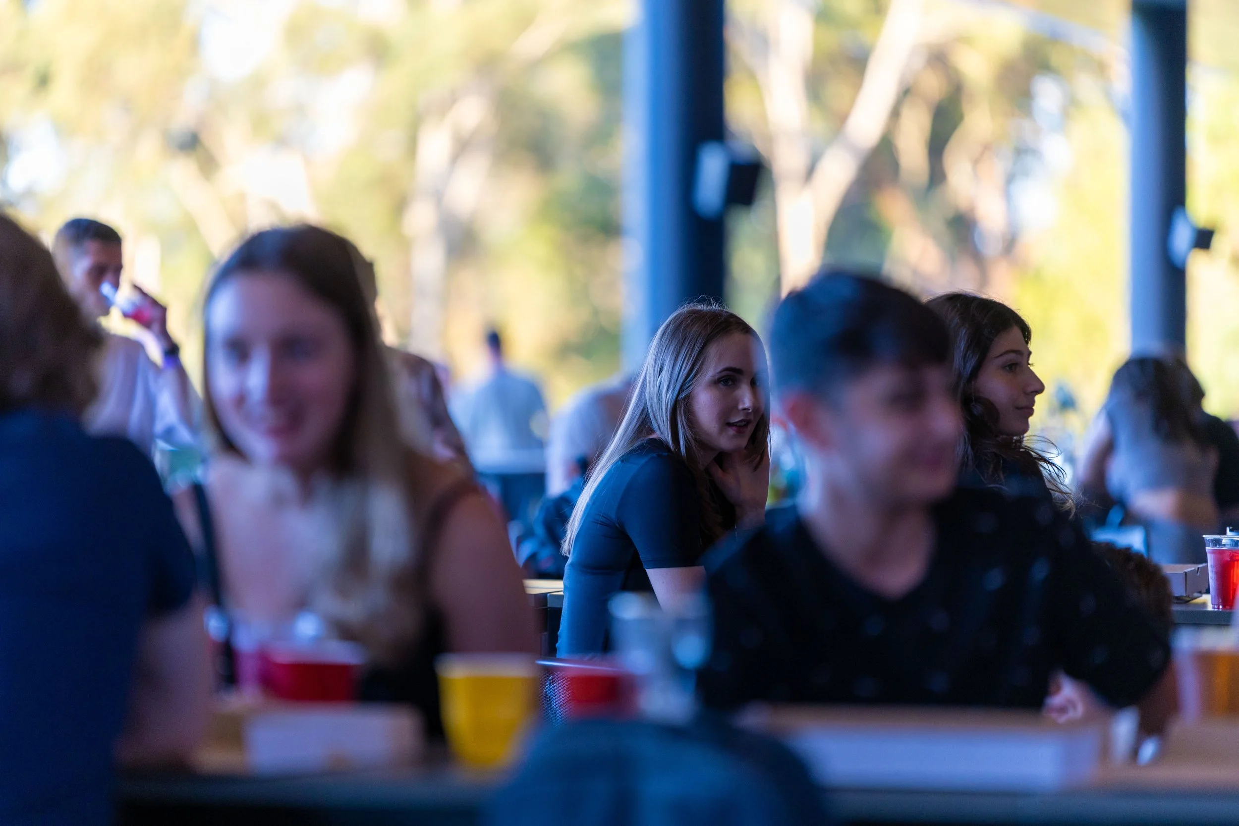People sitting at tables outdoors, engaged in conversation, with trees and blue structural beams in the background.