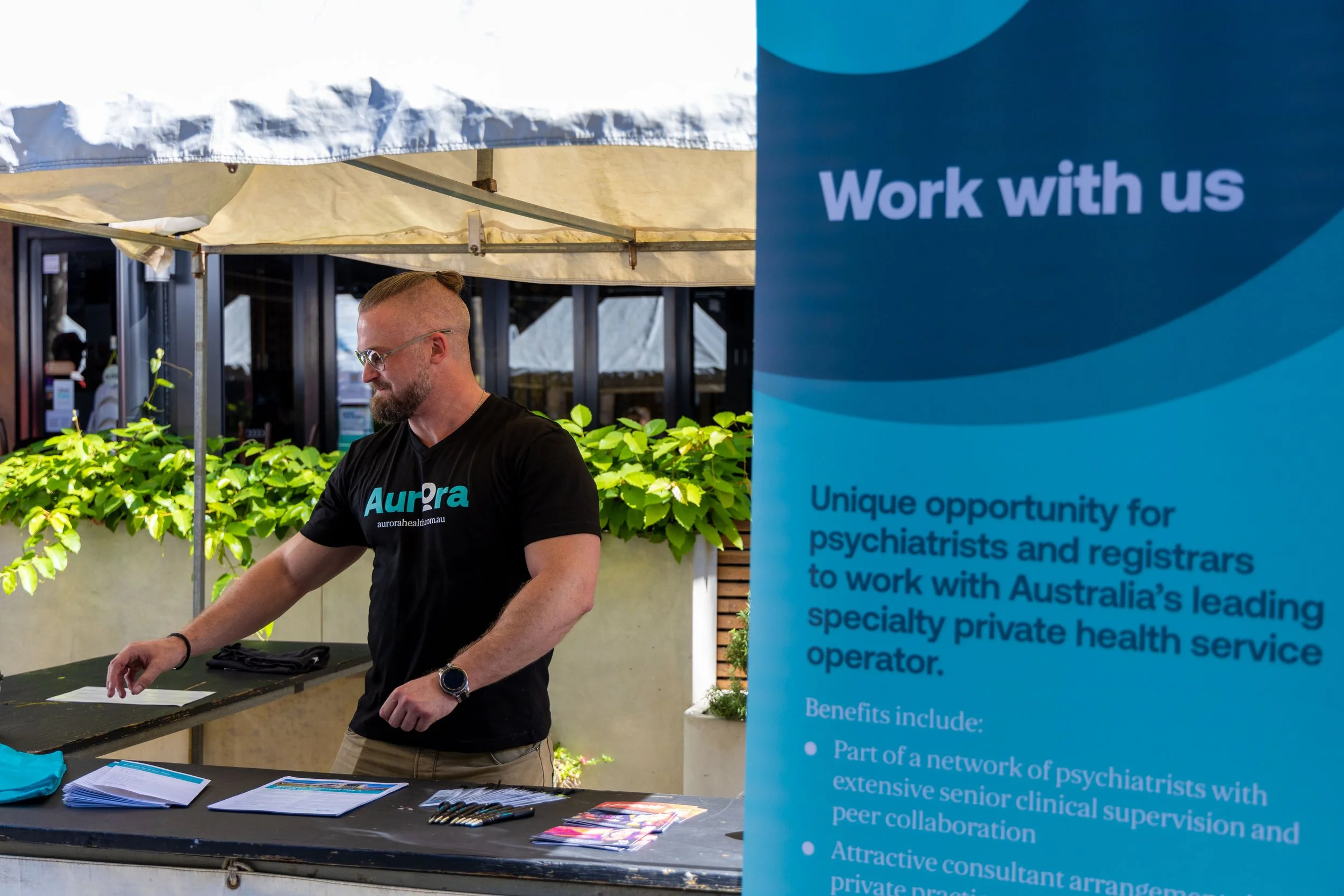 A man with a beard and glasses wearing a black t-shirt that says Aurora stands behind a table with brochures and pens at an outdoor job fair, next to a blue sign that reads 'Work with us' and describes opportunities for psychiatrists and registrars t